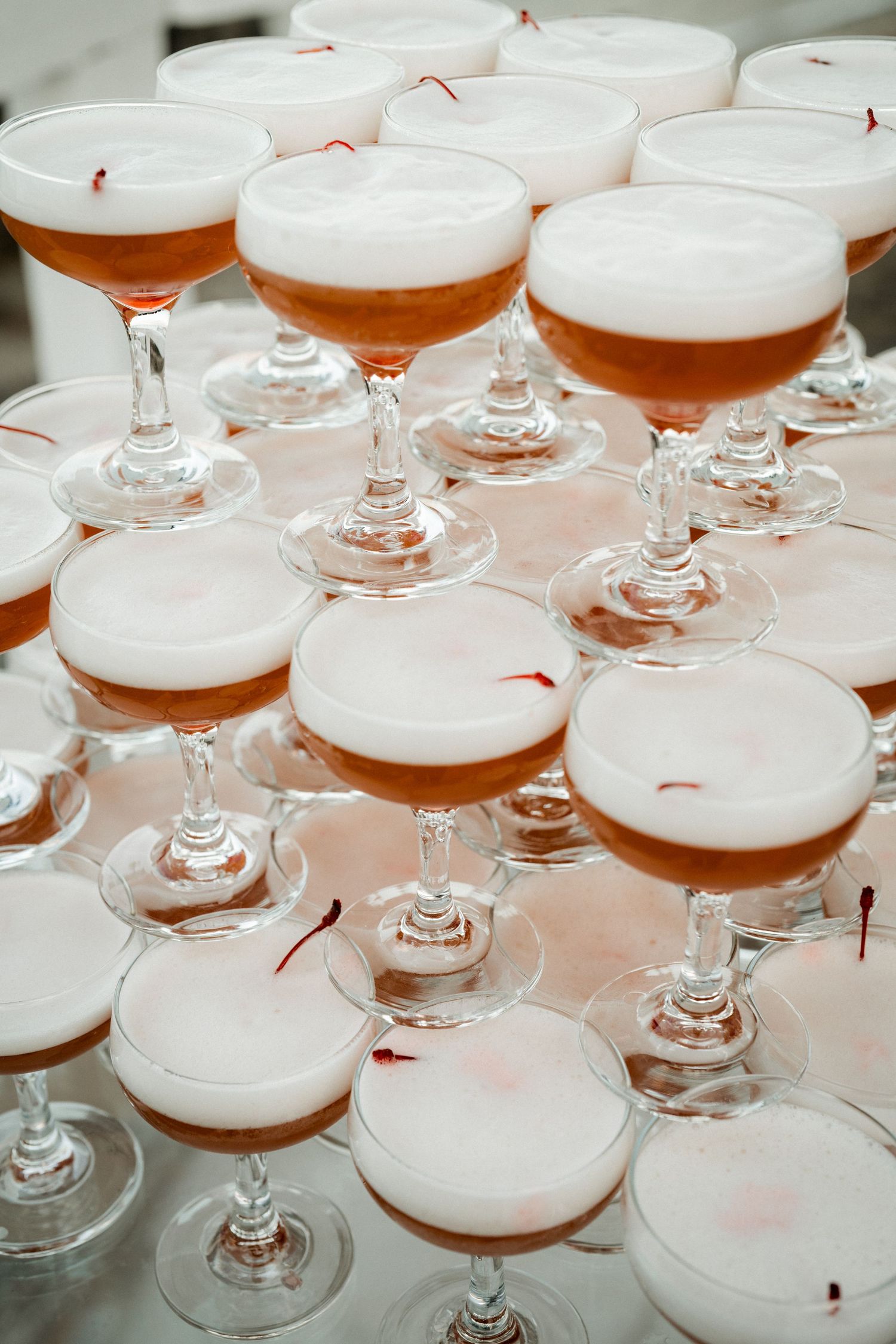 Multiple champagne coupes with frothy cocktails arranged on a reflective surface.