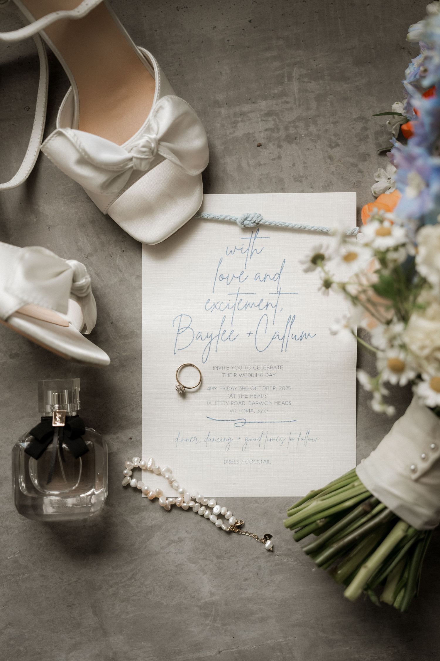 Wedding details including white shoes, floral bouquet, invitation cards, and rings arranged on a gray surface.