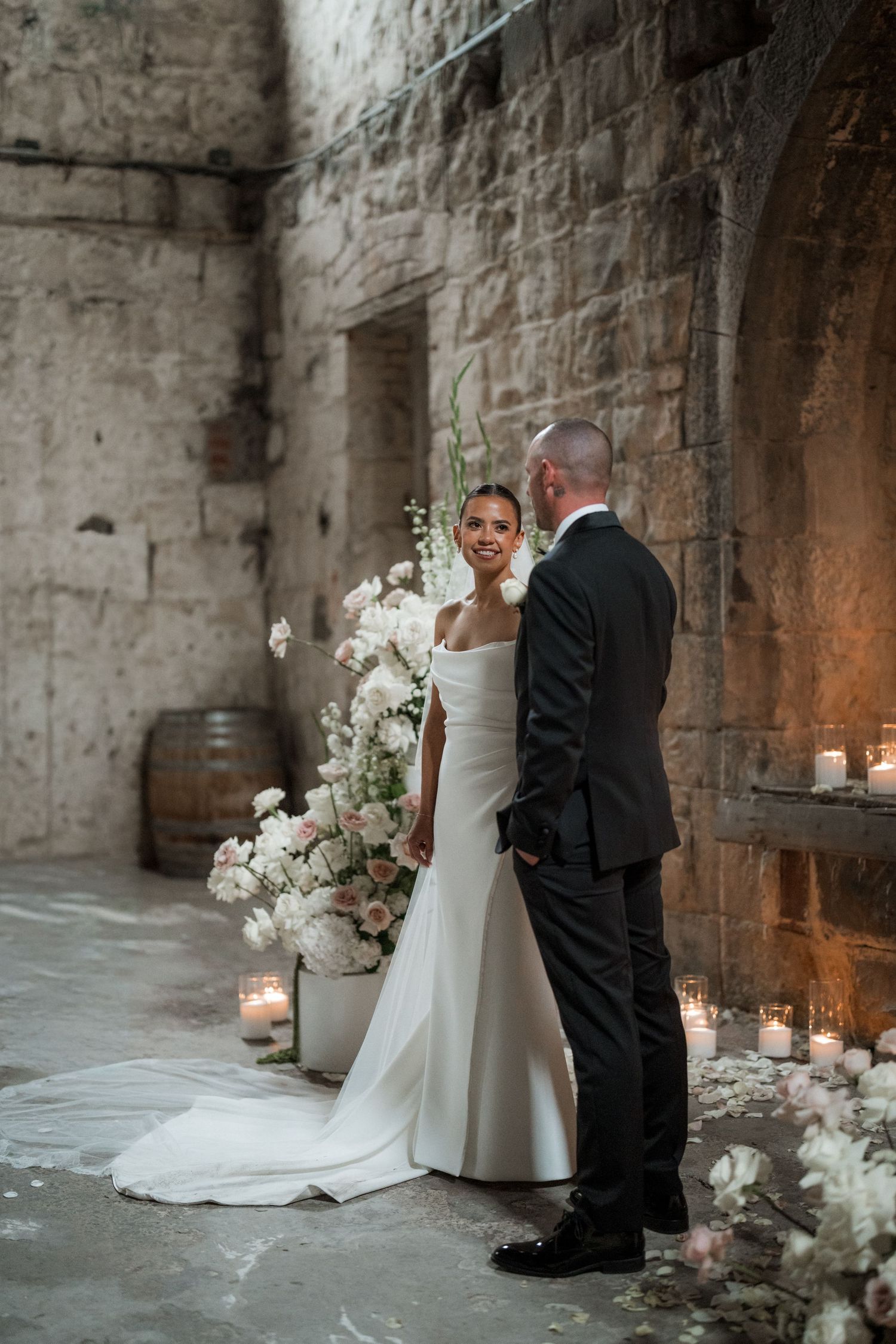 A couple poses for wedding photos in a rustic stone archway decorated with white floral arrangements.