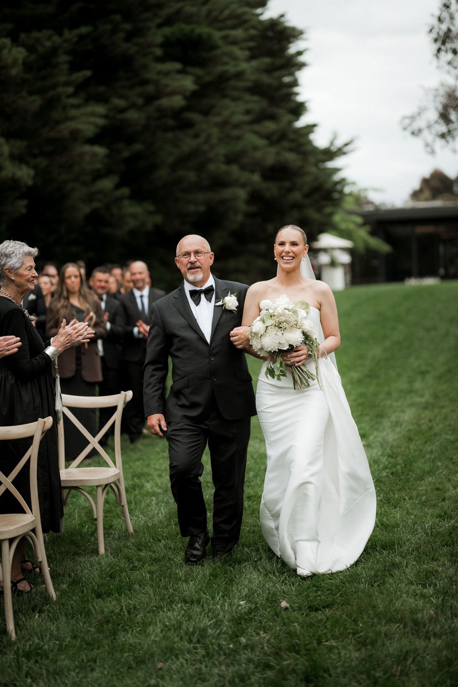 Bride in white gown walks down outdoor aisle with father in tuxedo, guests applauding on grass.