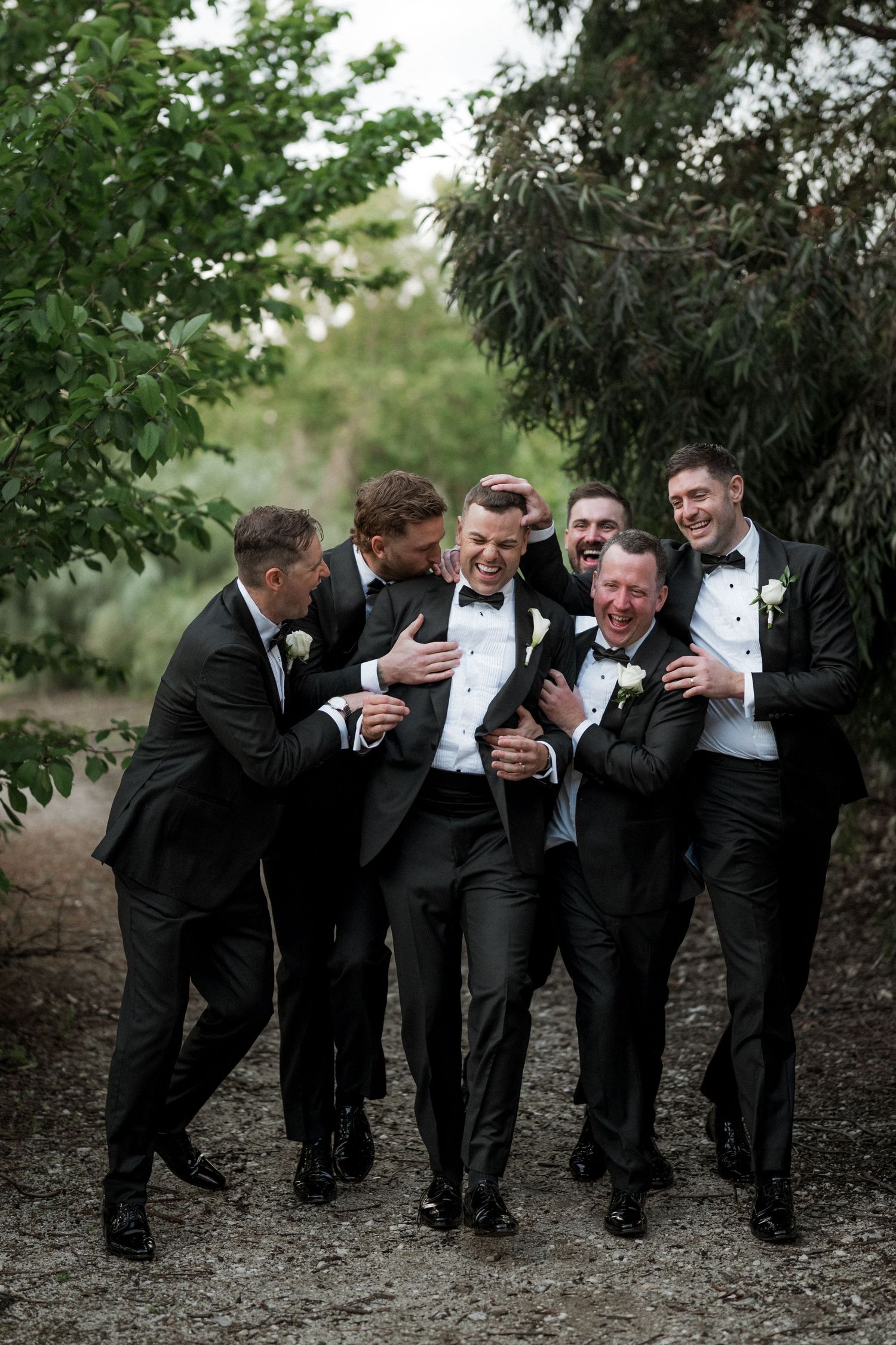 Groom and groomsmen laughing together in black tuxedos outdoors, sharing a joyful moment on the wedding day.