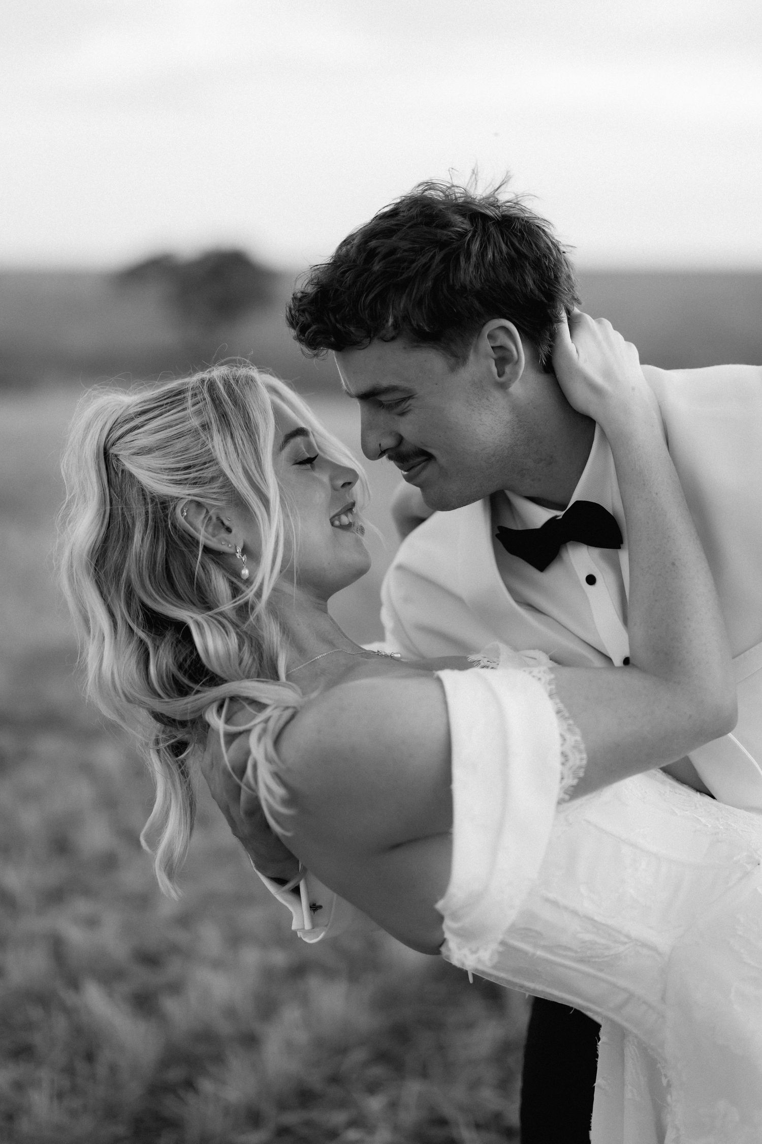 Black and white romantic photo of groom lifting bride in open field as they gaze at each other.