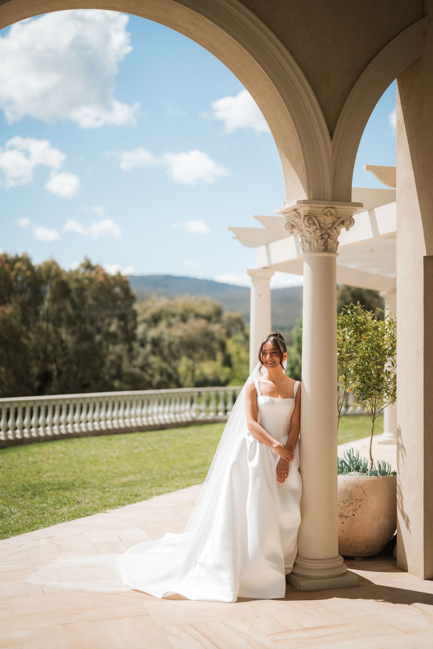 A bride in an elegant white satin gown and veil leans against a stone column on a sunlit terrace overlooking green hills.