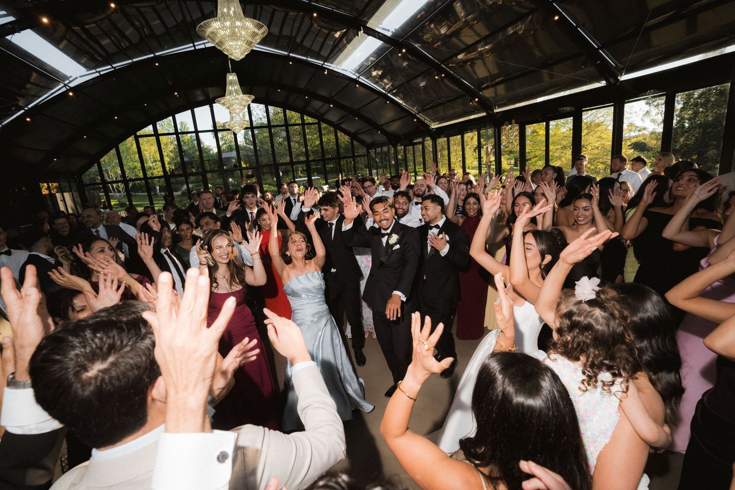 Formally dressed guests dance and raise their hands in celebration on a crowded dance floor inside a bright glass-walled event venue.