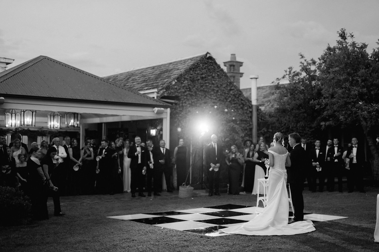 Bride and groom share first dance on checkered floor at elegant outdoor wedding reception in black and white photo.