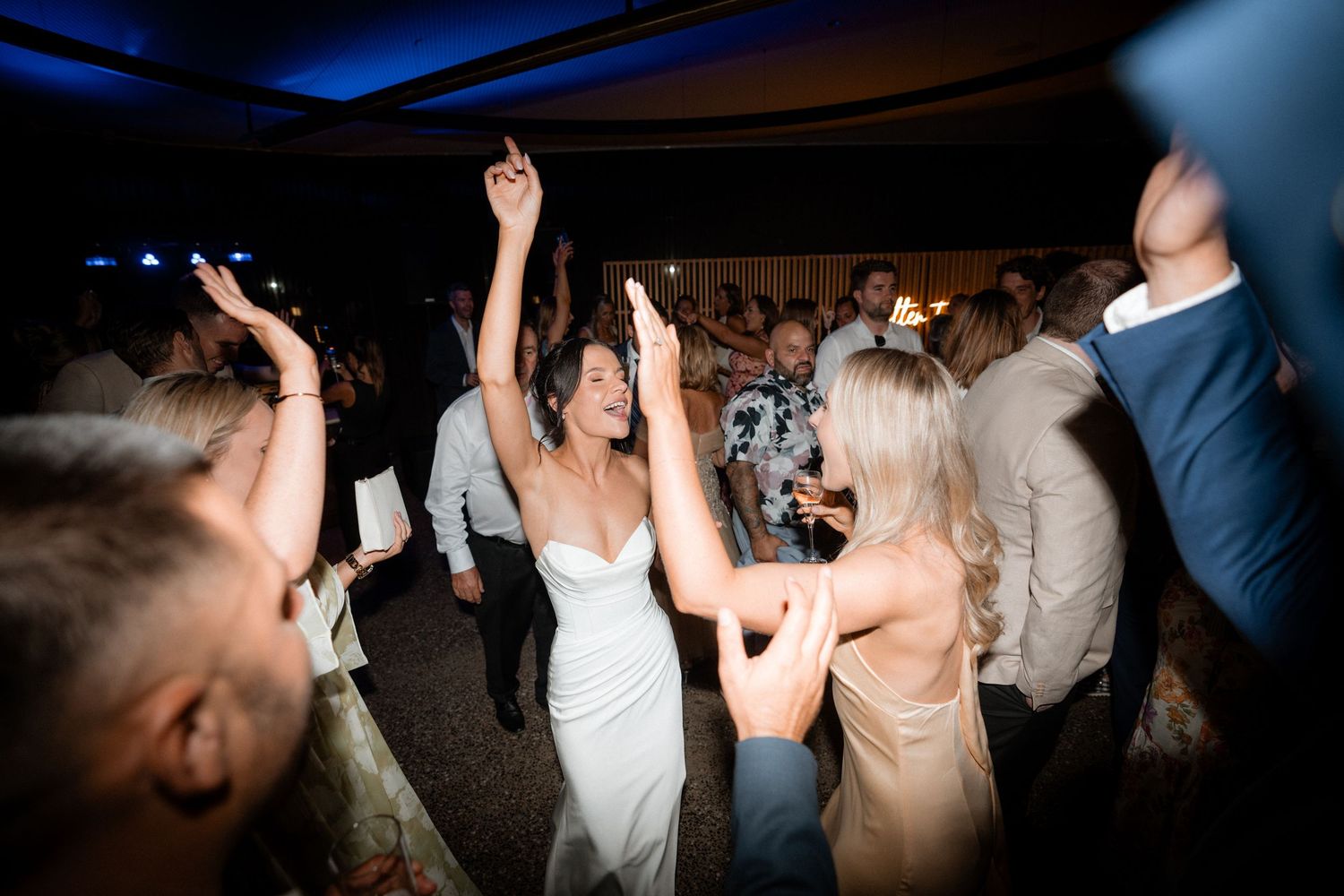 Wedding guests dance and celebrate together on a dark dance floor illuminated by blue lights during the reception party.