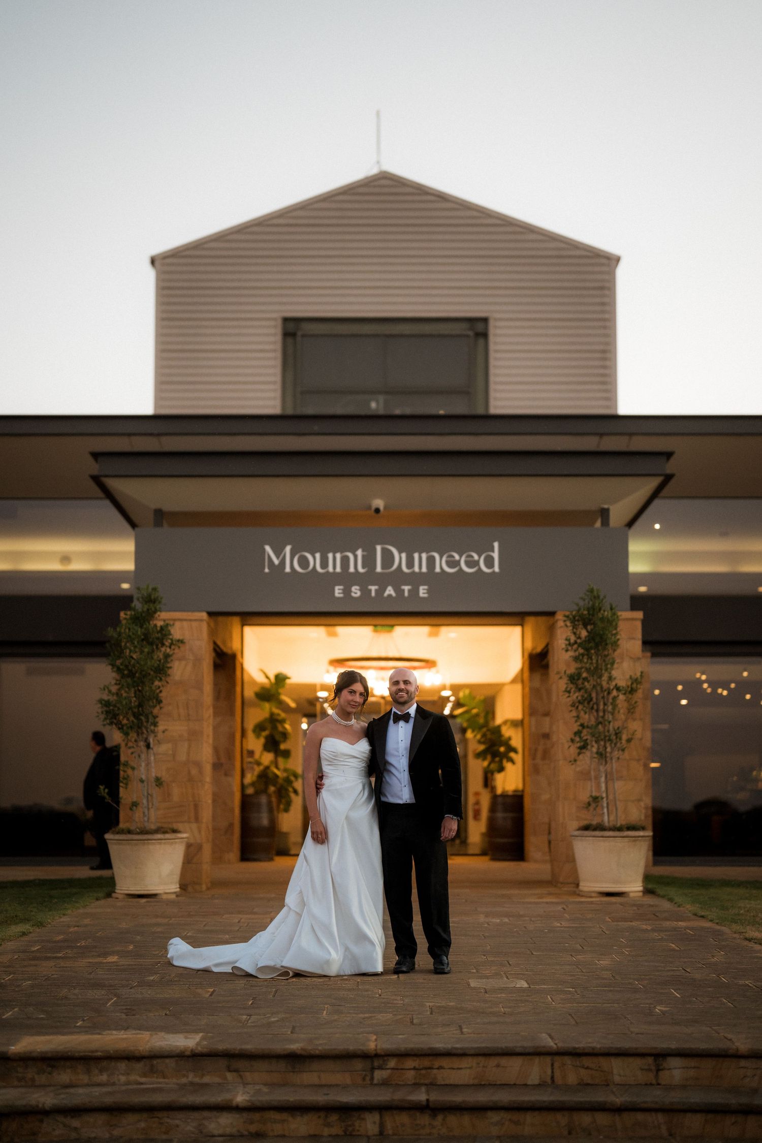 Wedding party poses outside Mount Duneed Estate winery entrance at dusk with illuminated signage.