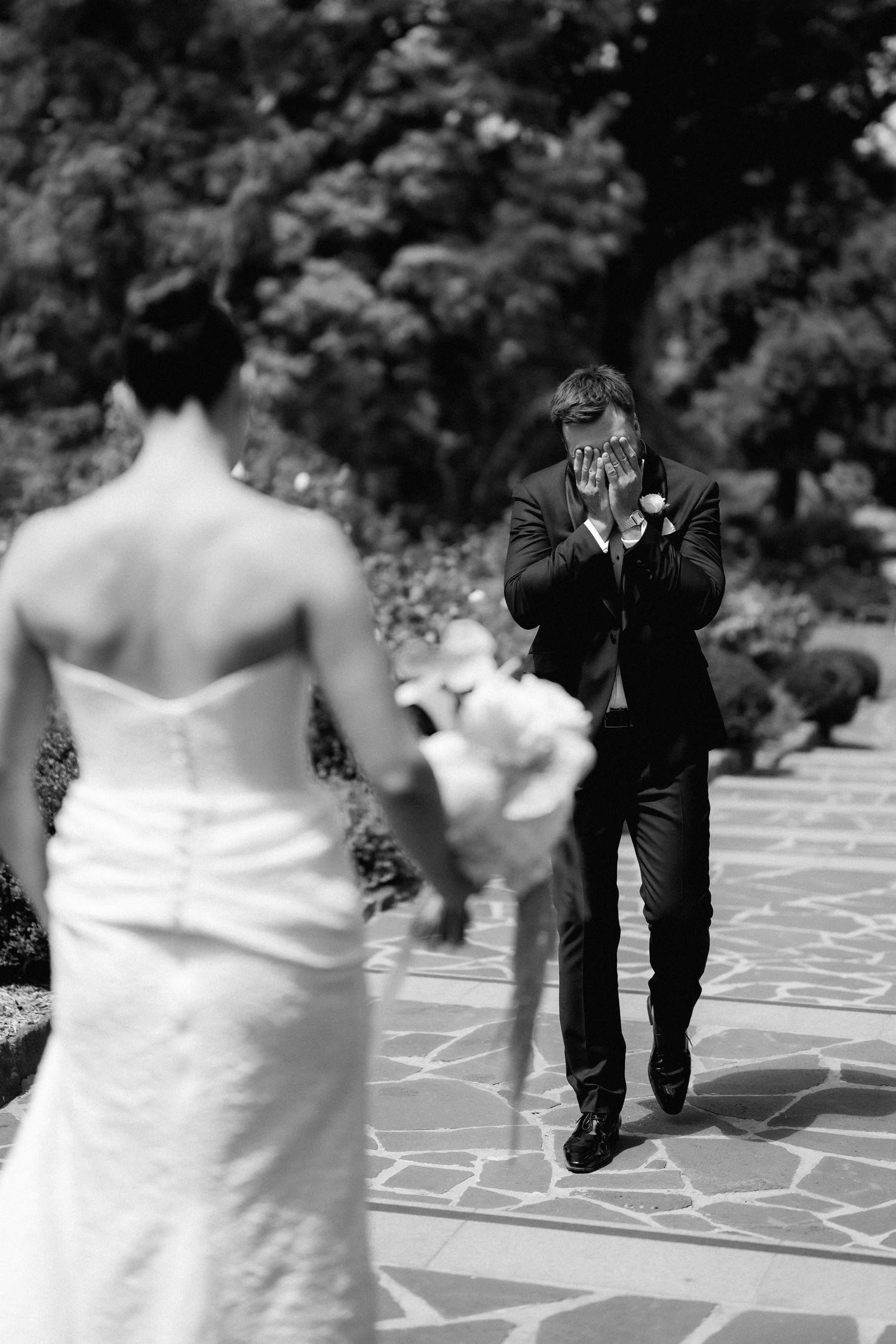 Emotional groom covering his face as bride approaches during first look at outdoor wedding.