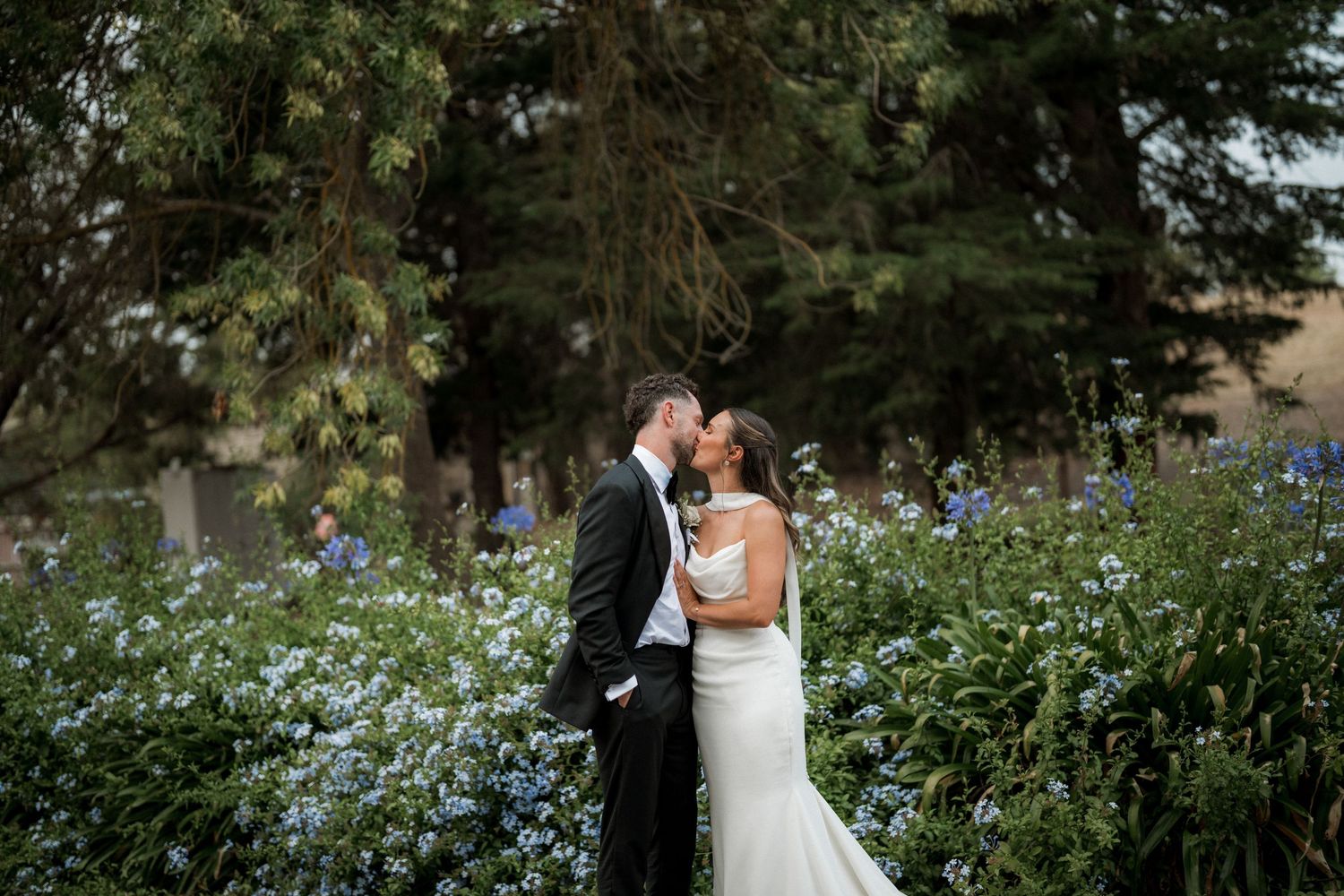 A romantic outdoor wedding scene with a couple embracing under tall evergreen trees.