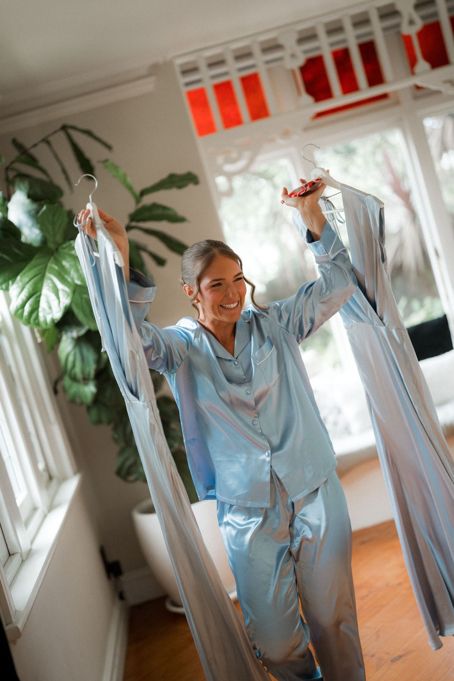 Person wearing light blue silk pajamas holds up clothing while standing in a sunlit room with houseplants.