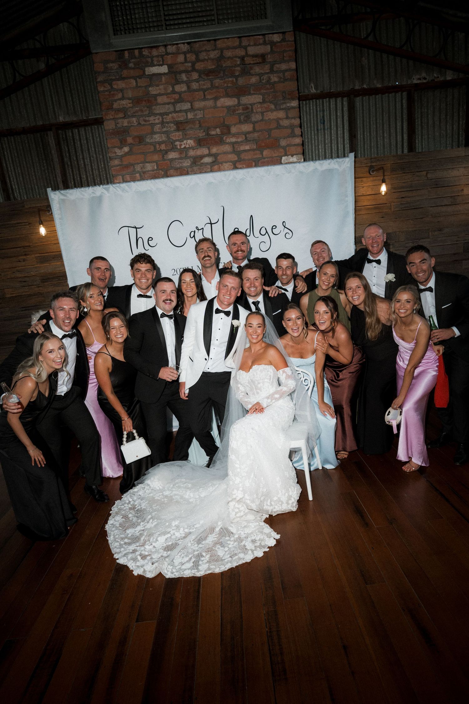 Wedding party poses together on a wooden dance floor in front of a brick wall at an indoor reception venue.