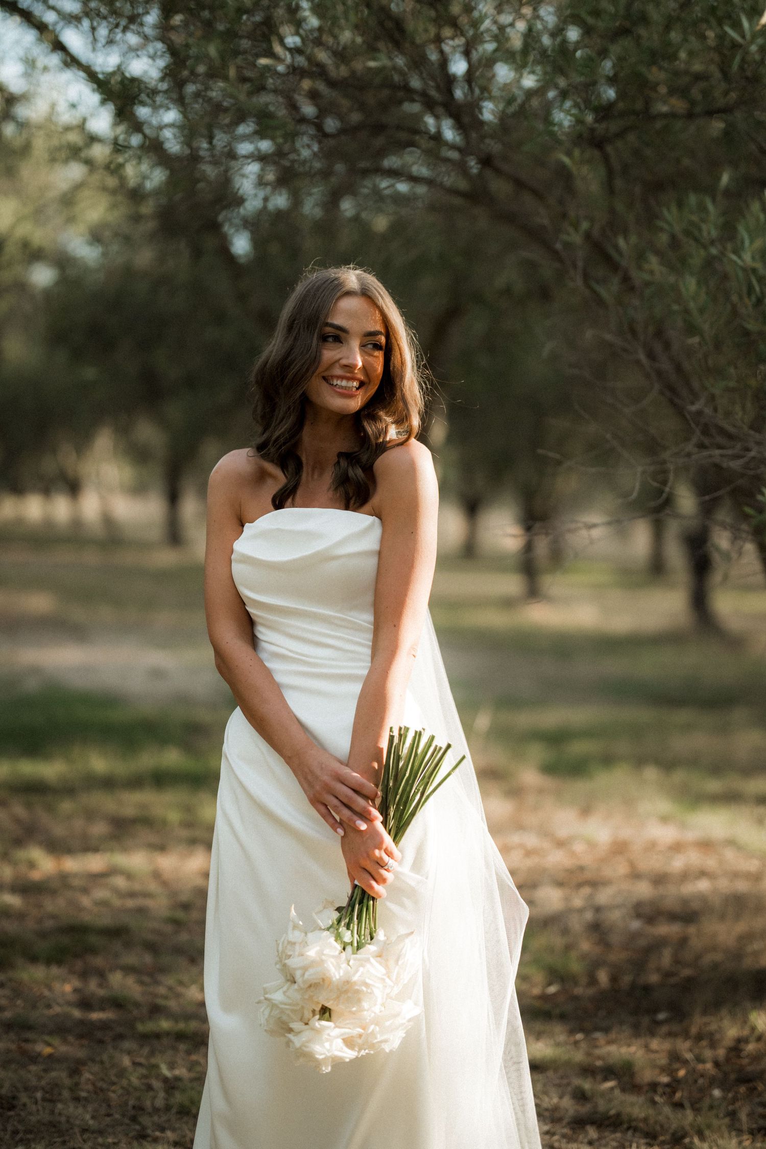 A bride in a white wedding dress holds a bouquet while standing in a sunlit orchard setting.
