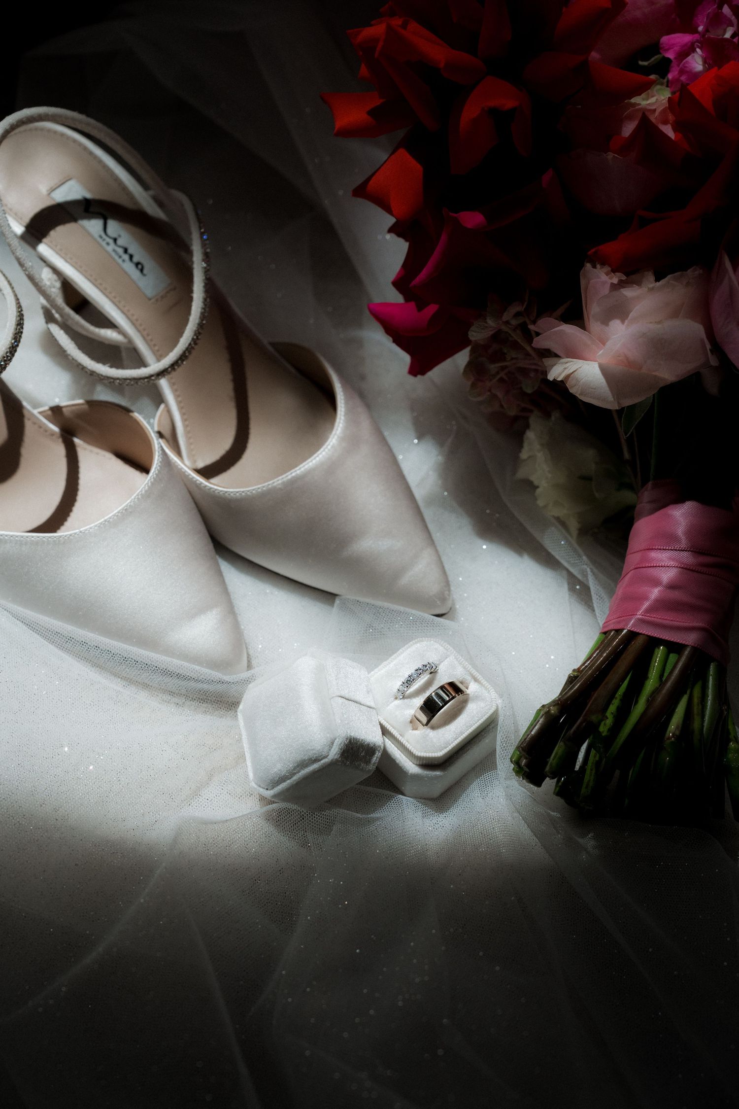 Elegant white bridal shoes and pink flowers arranged artistically in dramatic lighting.
