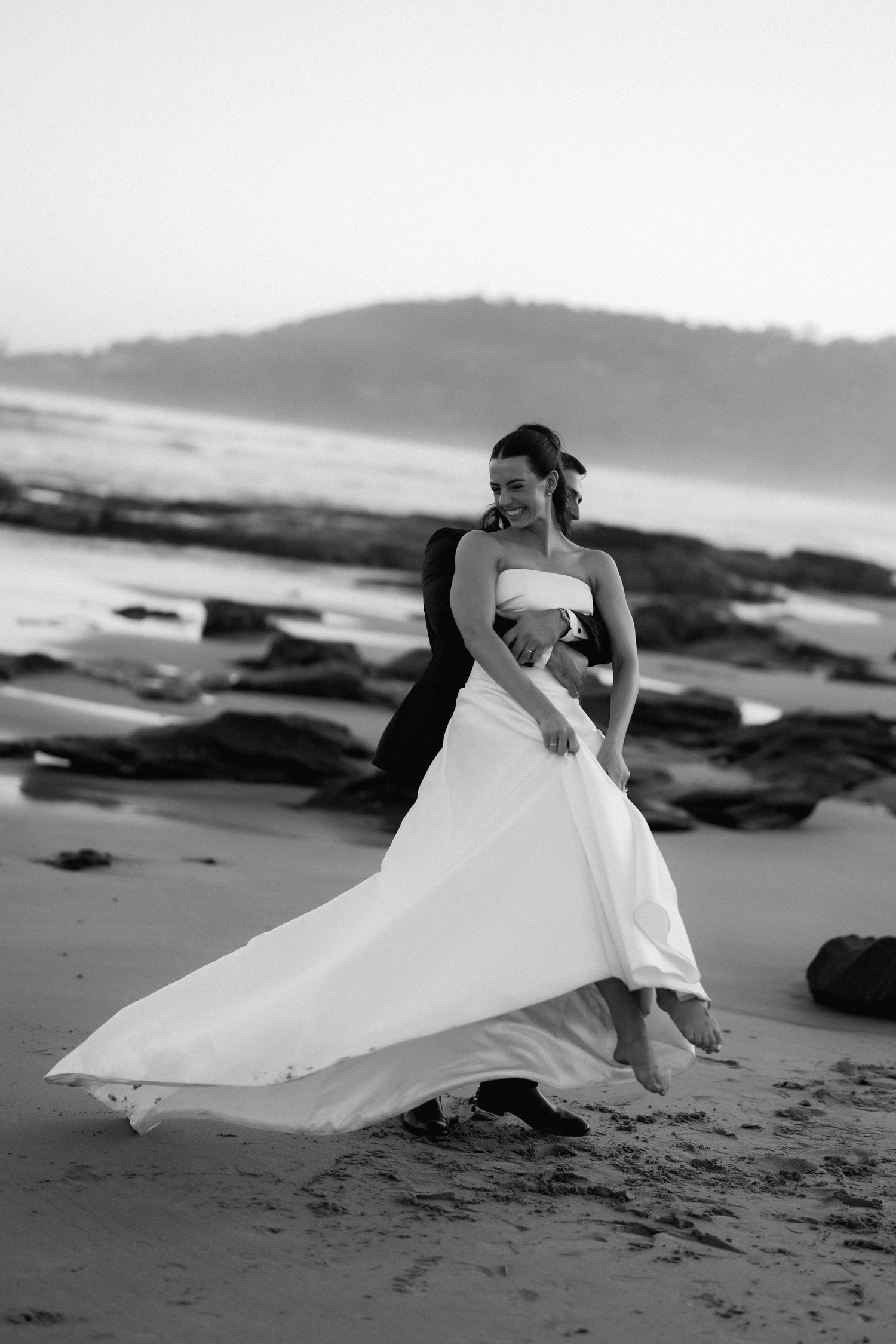 Black and white photo of couple in wedding attire posing dramatically on beach.