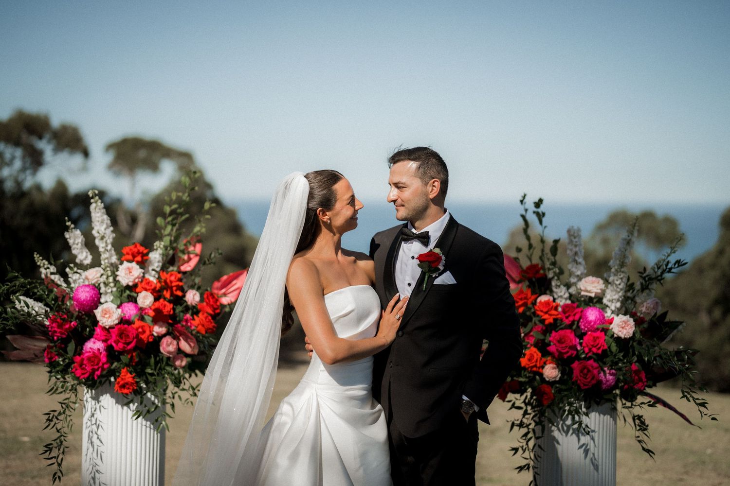 Outdoor wedding ceremony setup with vibrant pink and red floral arrangements against a coastal backdrop.