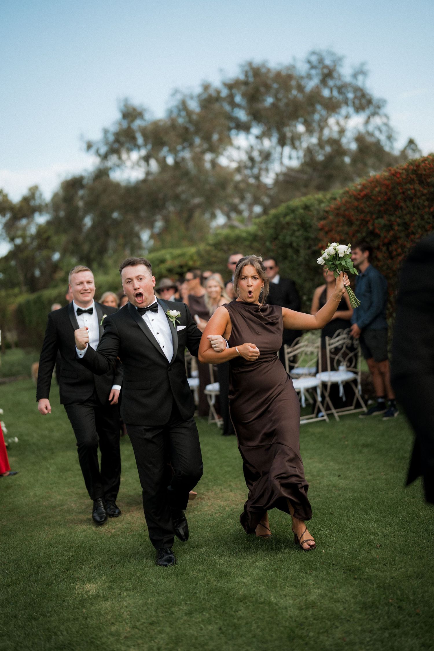 Wedding party members dancing joyfully on grass during outdoor celebration.