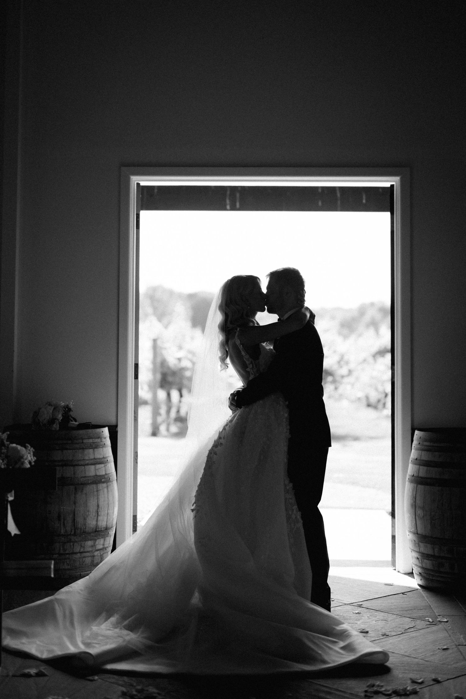 Silhouetted newlyweds share an intimate moment in a doorway framed by wine barrels.