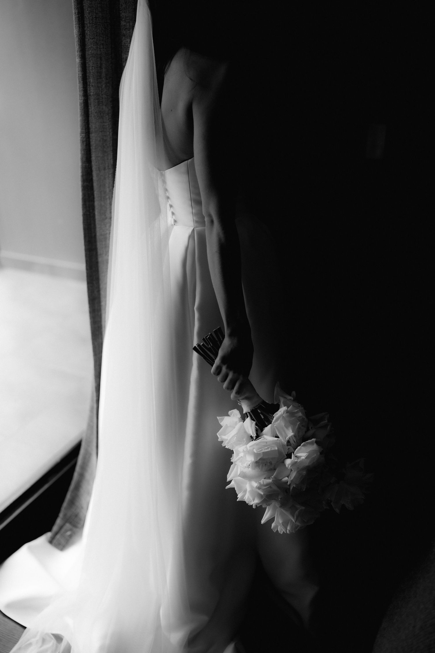 Artistic black and white detail shot of wedding dress and bouquet near window.