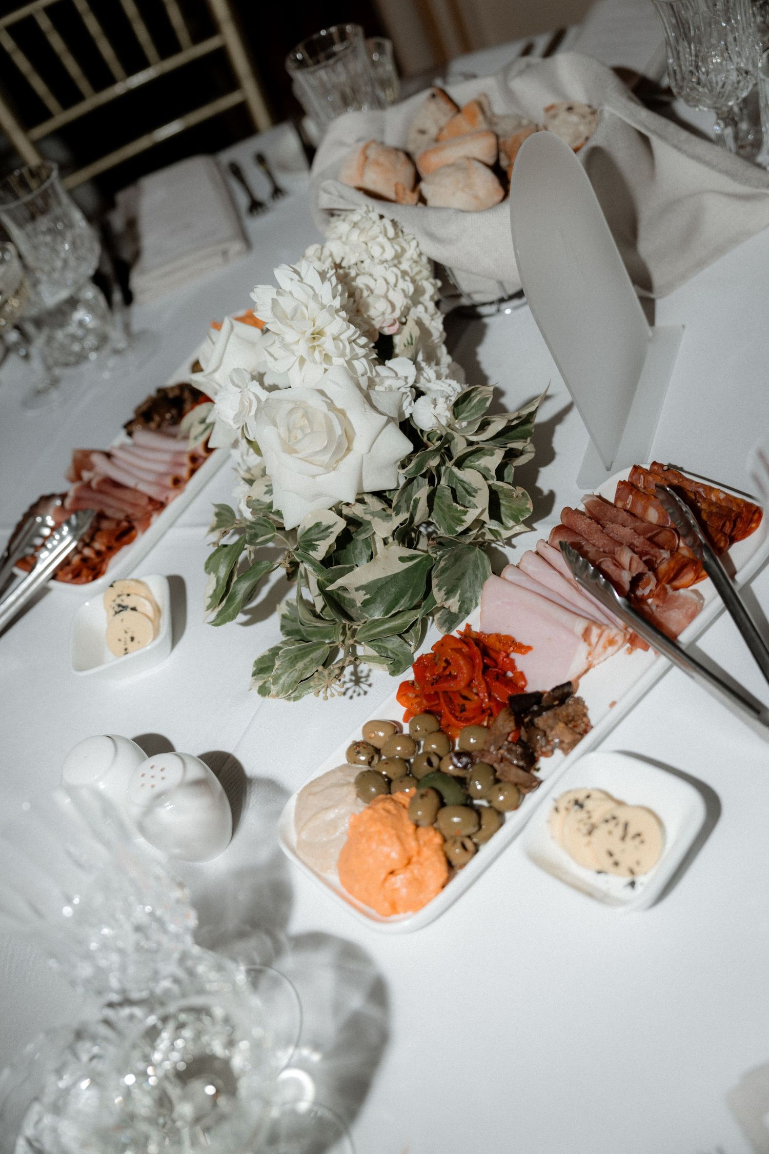 Elegant table setting with white roses, fresh greens and charcuterie appetizers on a white tablecloth.