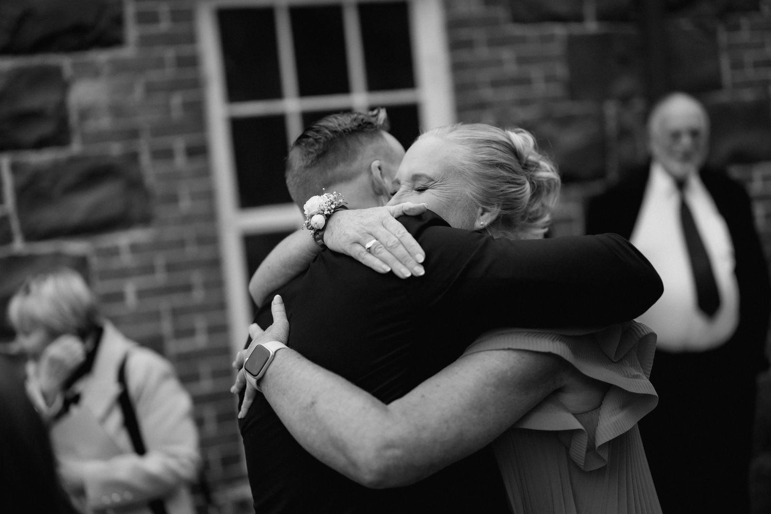 A black and white photograph of two people embracing in an intimate hug outside a brick building.