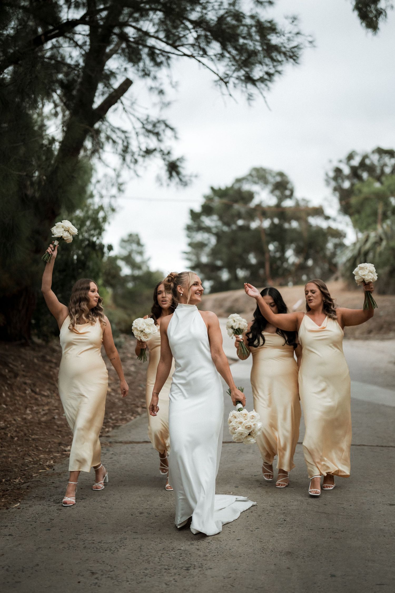 Wedding party walking down path with women in yellow dresses holding bouquets and smiling.