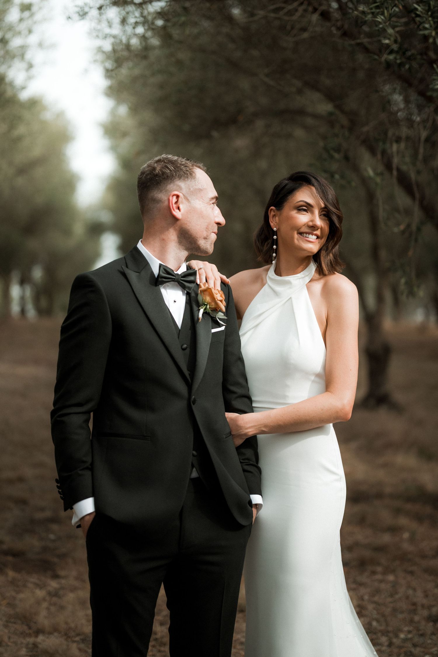 A happy couple shares a joyful moment together in formal wedding attire on a tree-lined path.