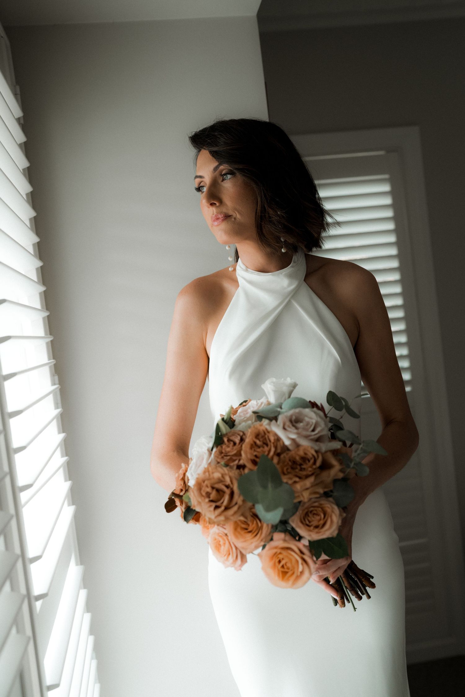 Beautiful bridal portrait in elegant white halter dress holding a romantic bouquet of peach roses by window light.