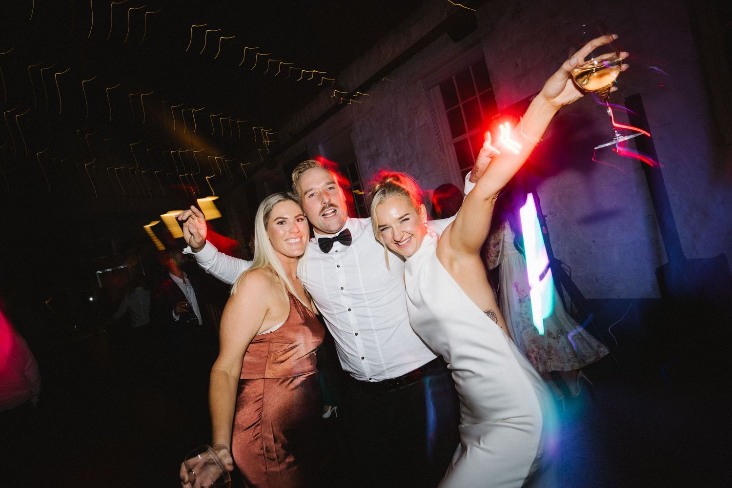 People celebrating and dancing energetically at a nighttime party with colorful lights in the background.
