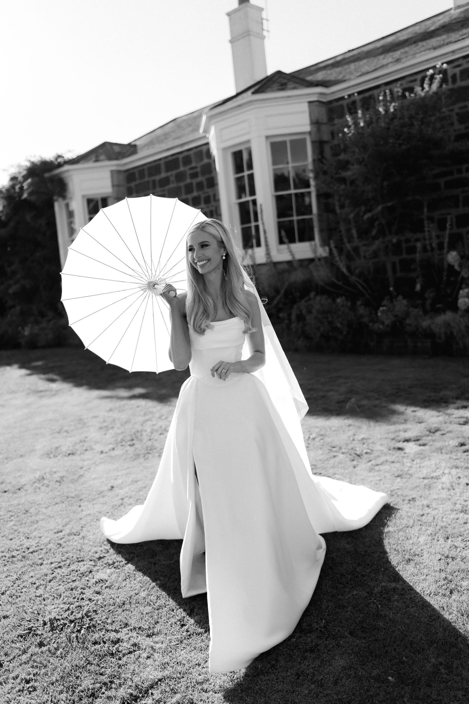 Bride in elegant white wedding gown poses with white parasol umbrella in black and white outdoor photograph.