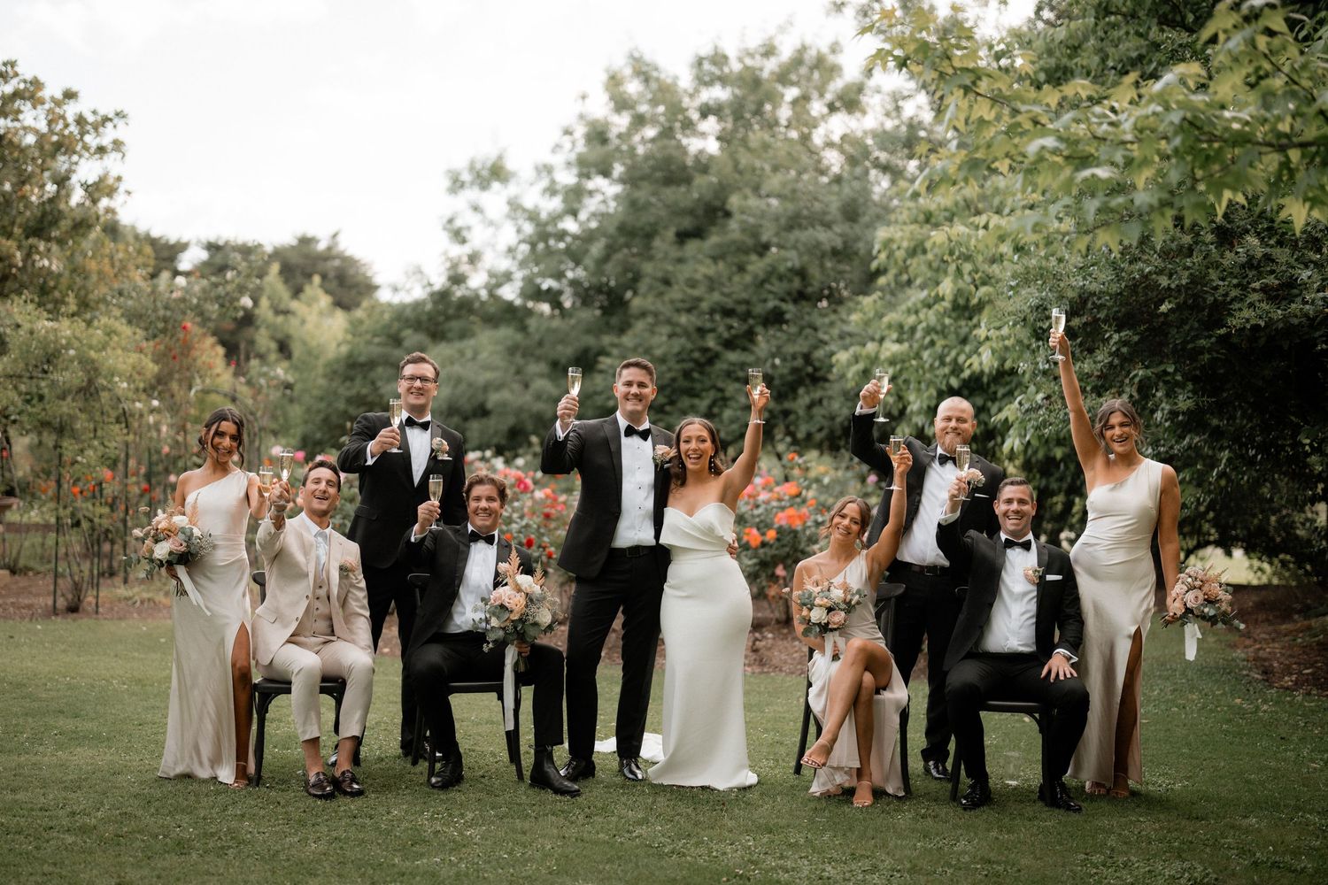 Wedding party members celebrate outdoors with raised arms in a joyful group pose on a grassy lawn.