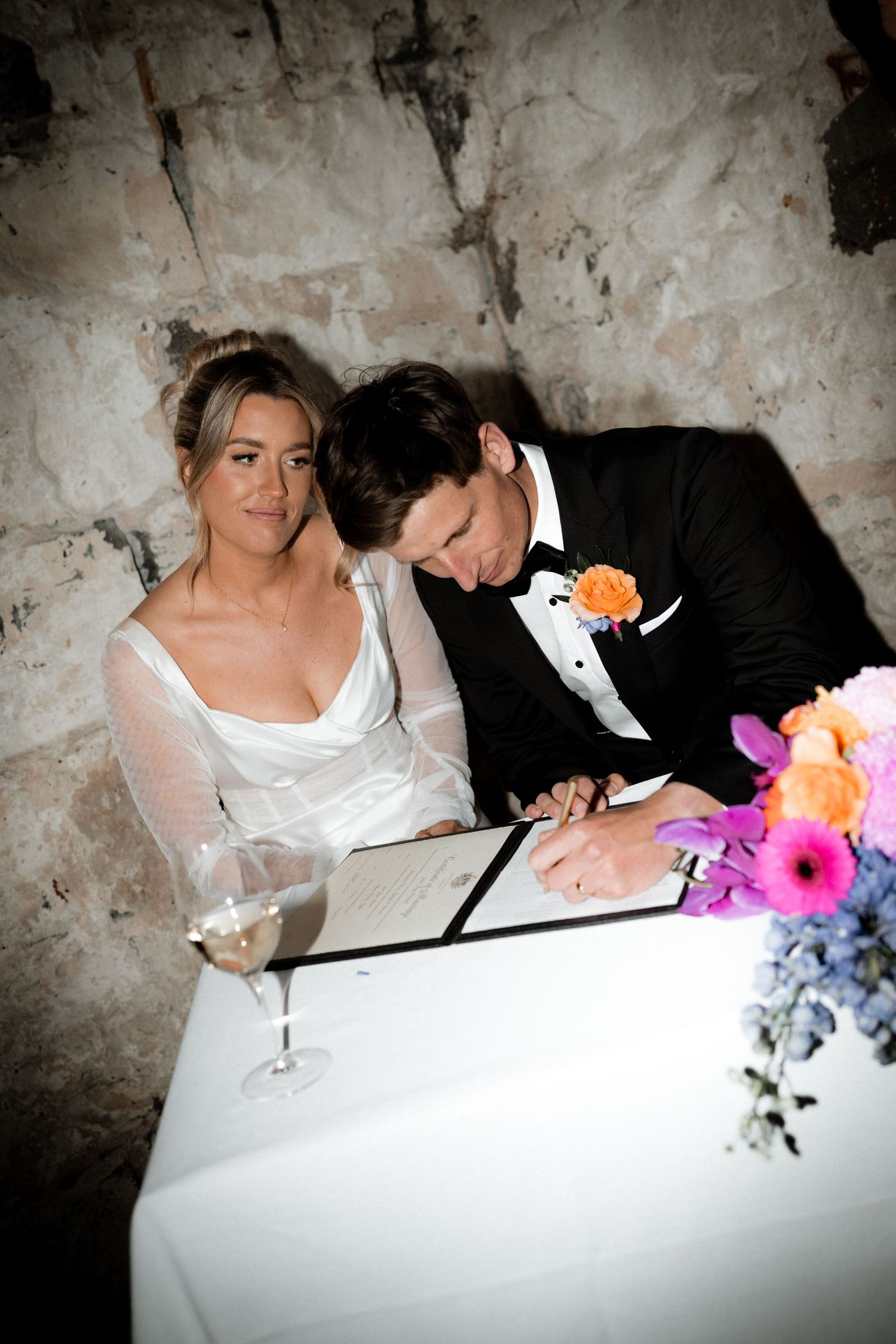 A bride and groom sign their marriage certificate at a stone-walled venue with colorful floral decorations and wine glass.