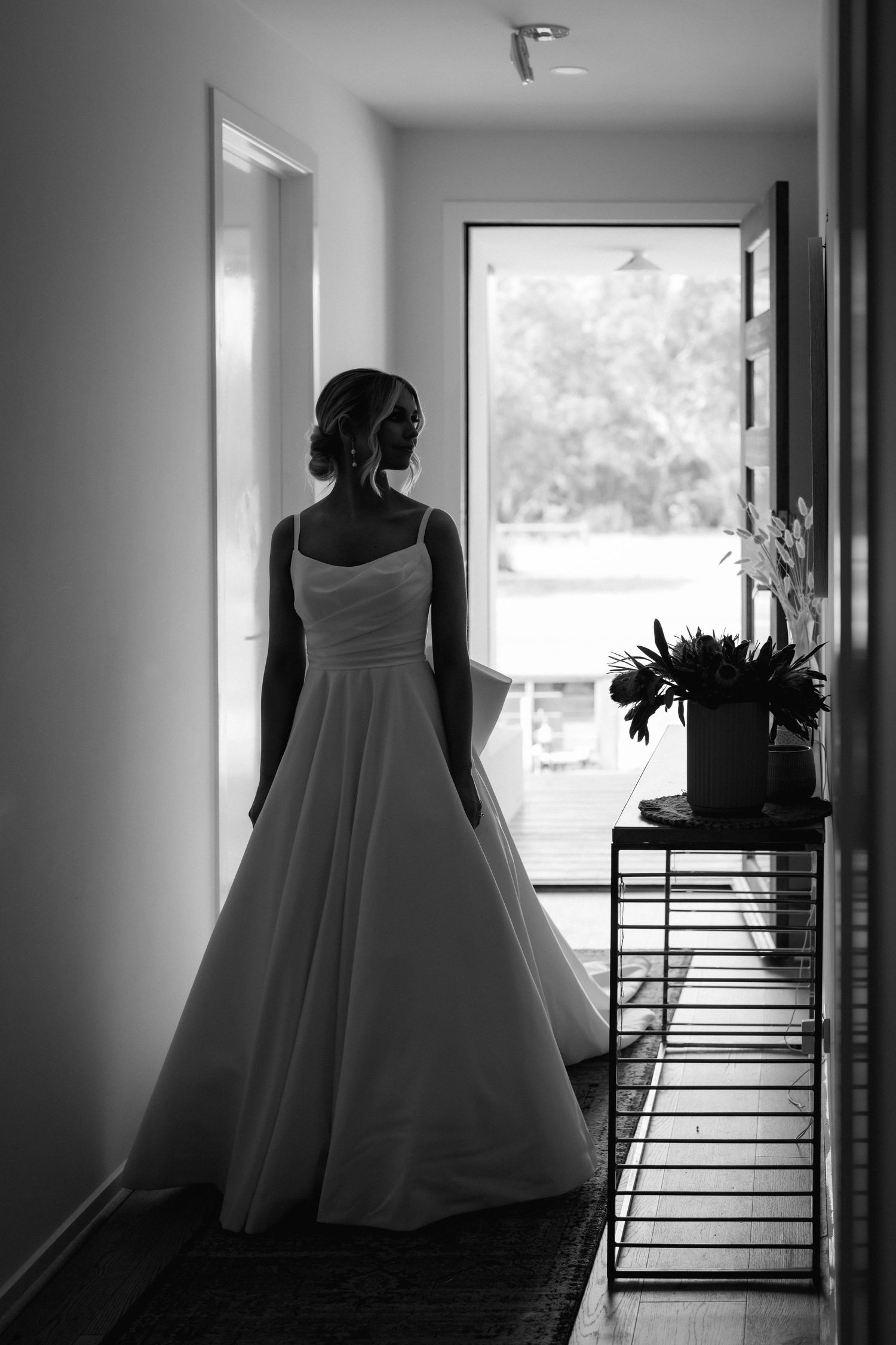 A black and white photo of a person in a wedding dress standing by a window in silhouette.
