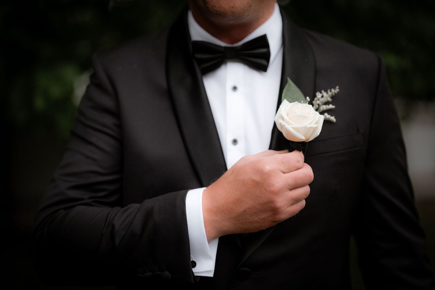 A close-up shot of a groom in a black tuxedo and bow tie adjusting his boutonniere flower.