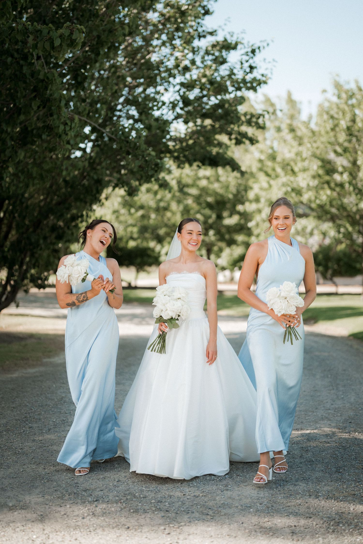 Wedding party in blue and white dresses share candid moments on a sunny path.