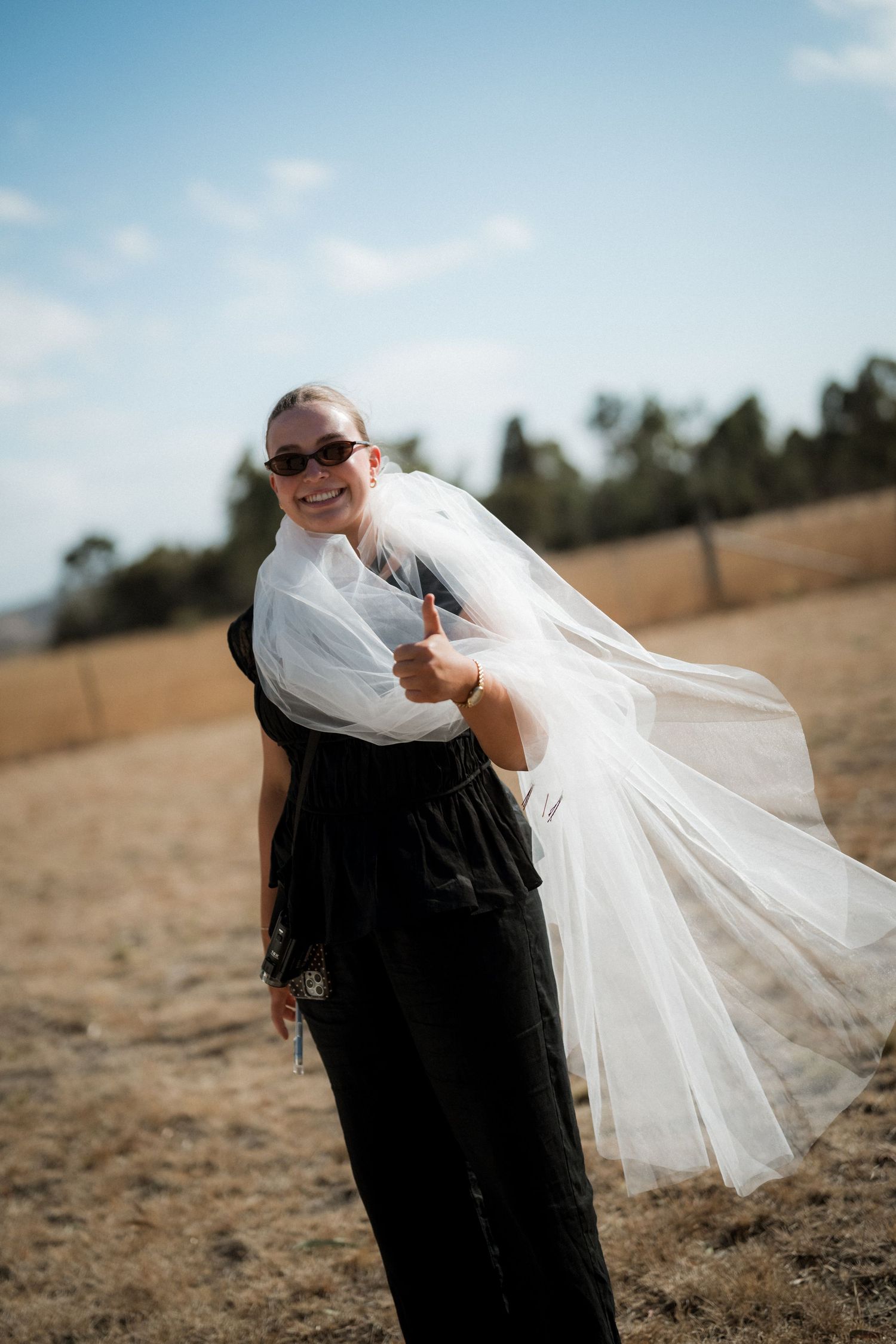 A flowing white wedding veil billows dramatically in the wind against a sunny outdoor backdrop.