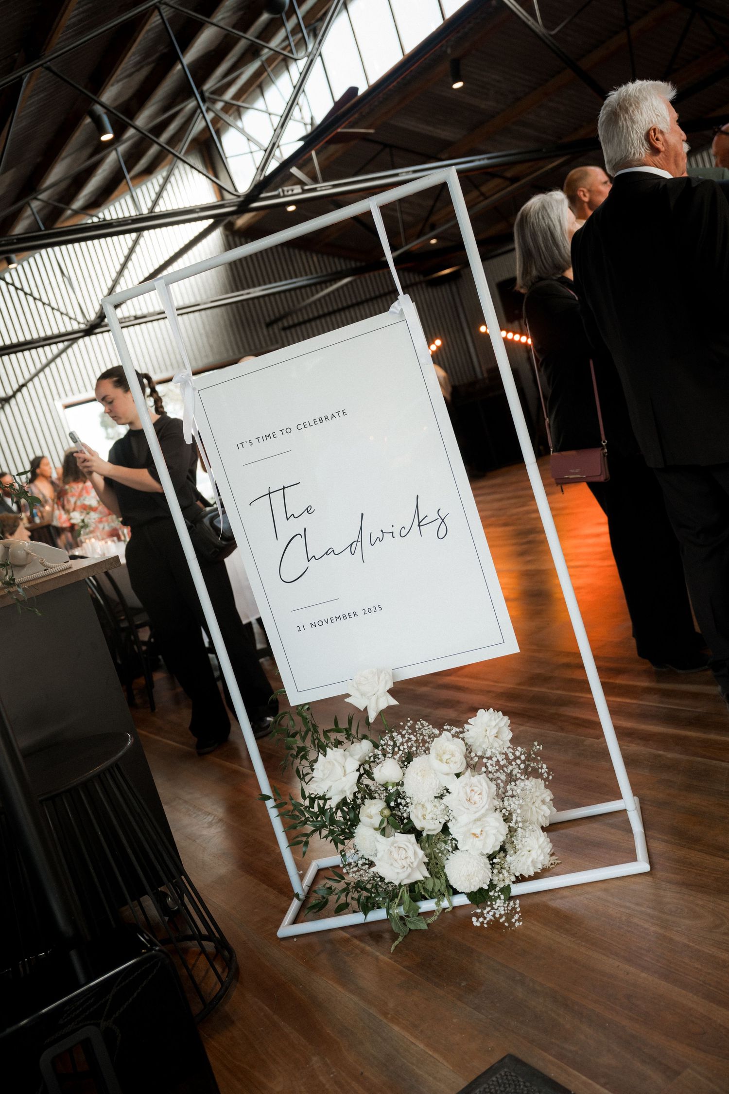 White floral wedding decor display with modern frame signage on wooden floor at indoor venue.