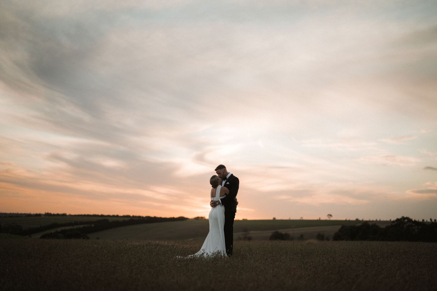 Silhouettes of wedding couple stand against a dramatic sunset sky over an open field during golden hour.