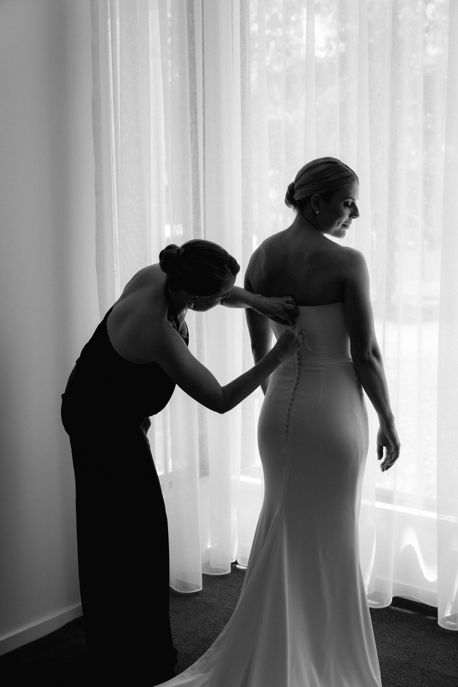 A silhouette of someone helping adjust a wedding dress in front of sheer white curtains.