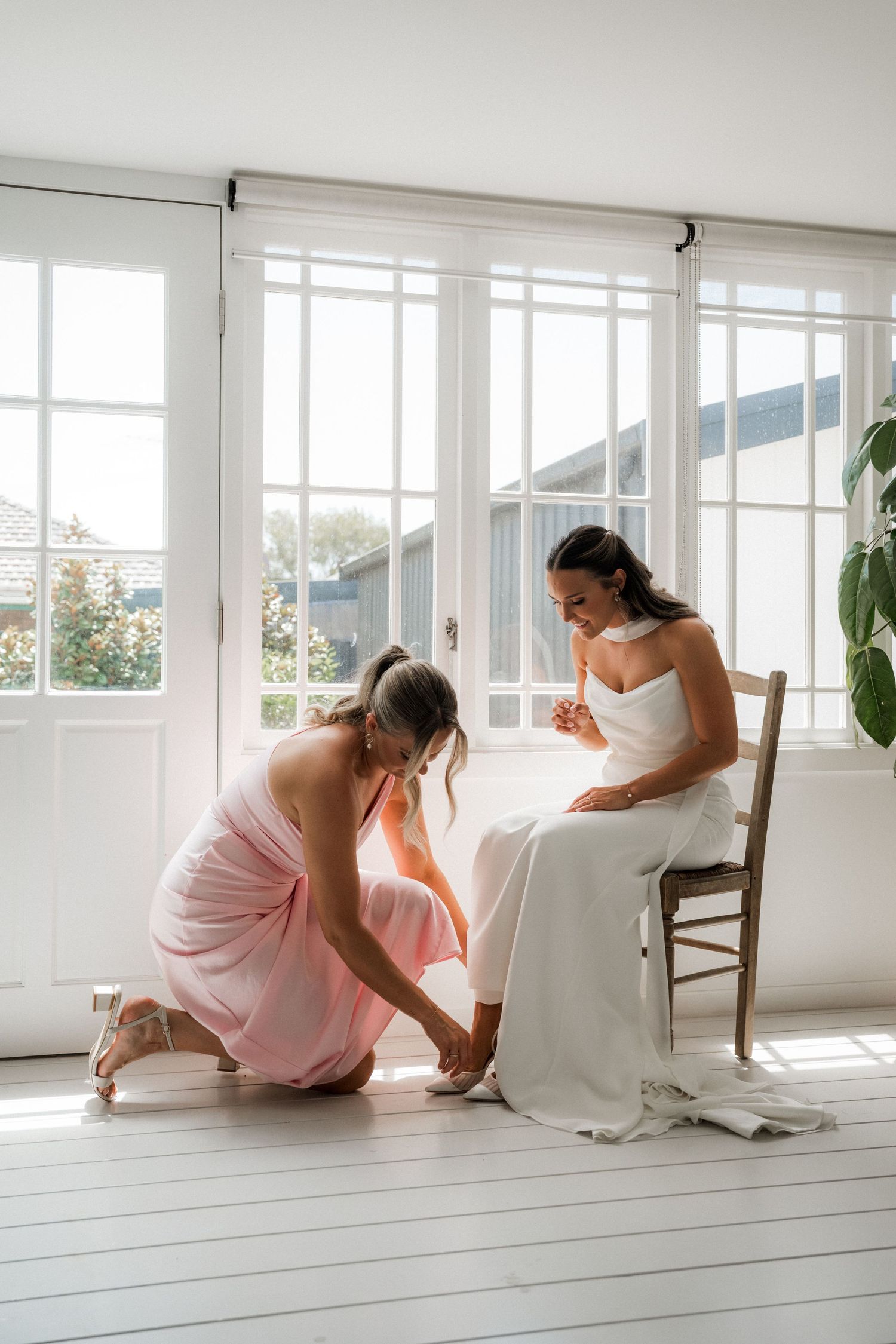 Bridesmaids in pink dresses relax on white window bench during wedding preparations.