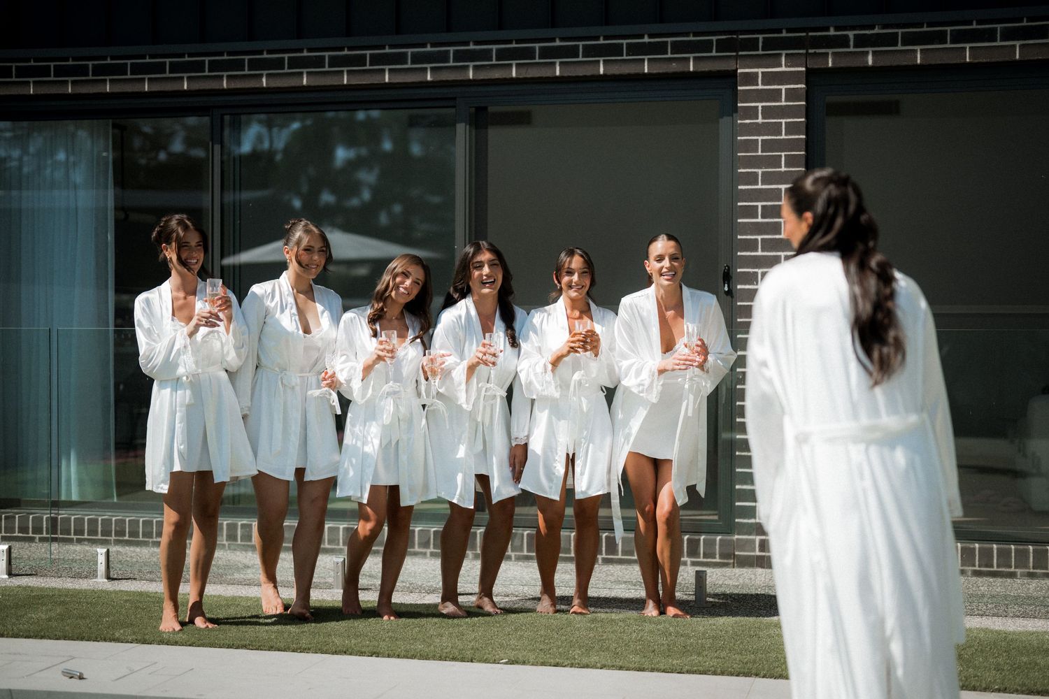Bridesmaids in white robes laugh together while getting ready for wedding day outside brick building.