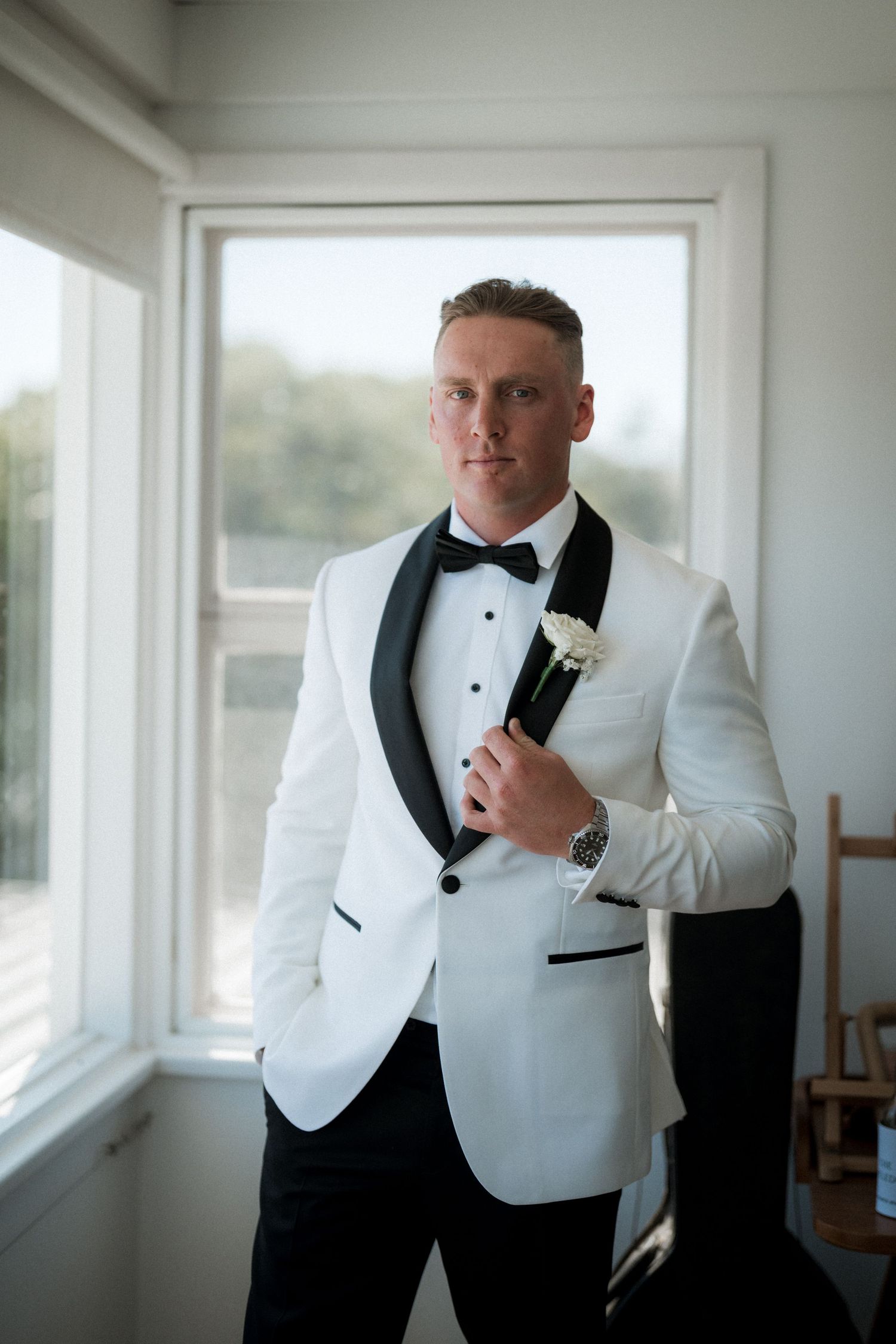 A groom adjusts his white tuxedo jacket with black lapels in a sunlit room before the wedding ceremony.