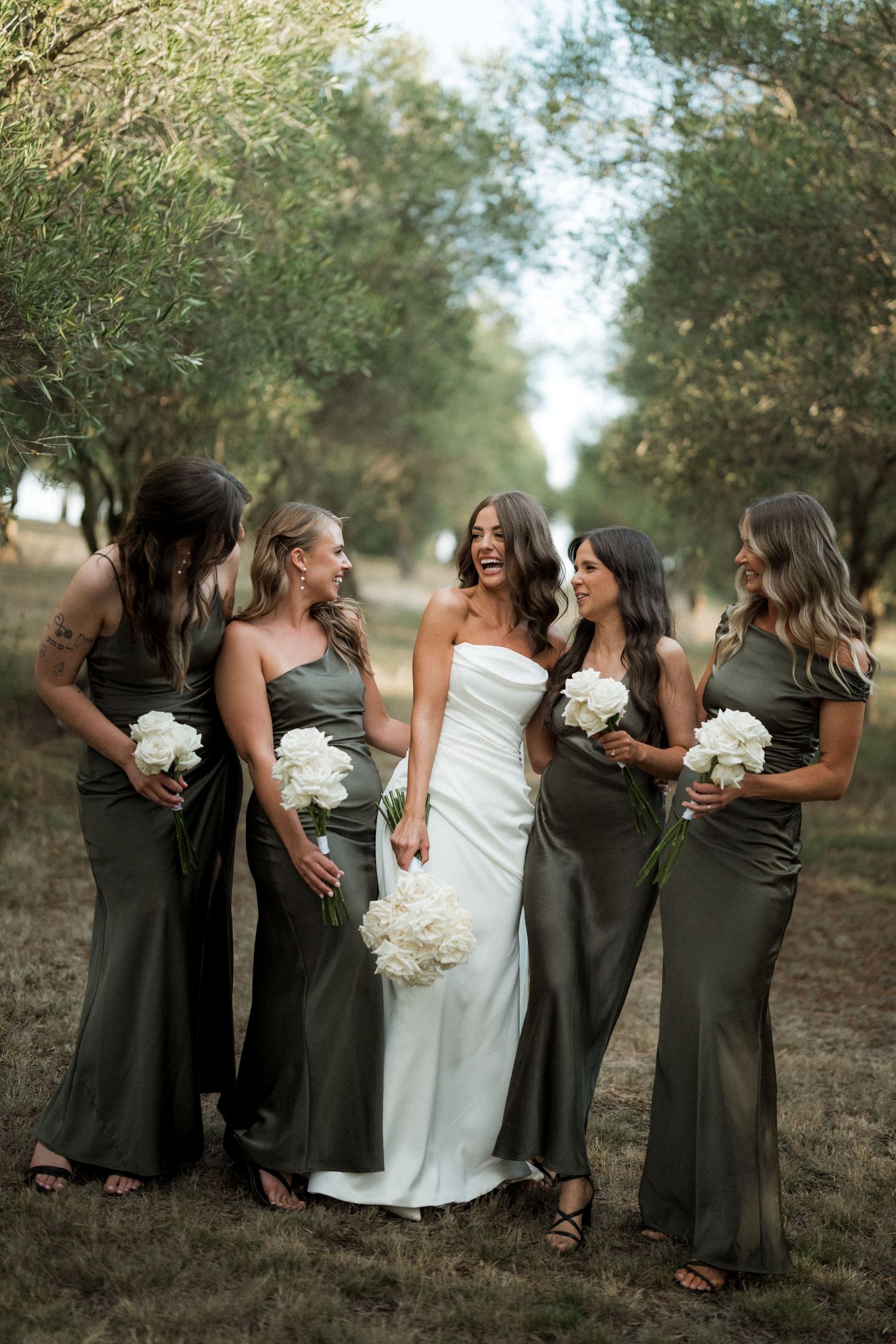 A bridal party in olive green dresses and white gown poses playfully in an outdoor orchard setting.