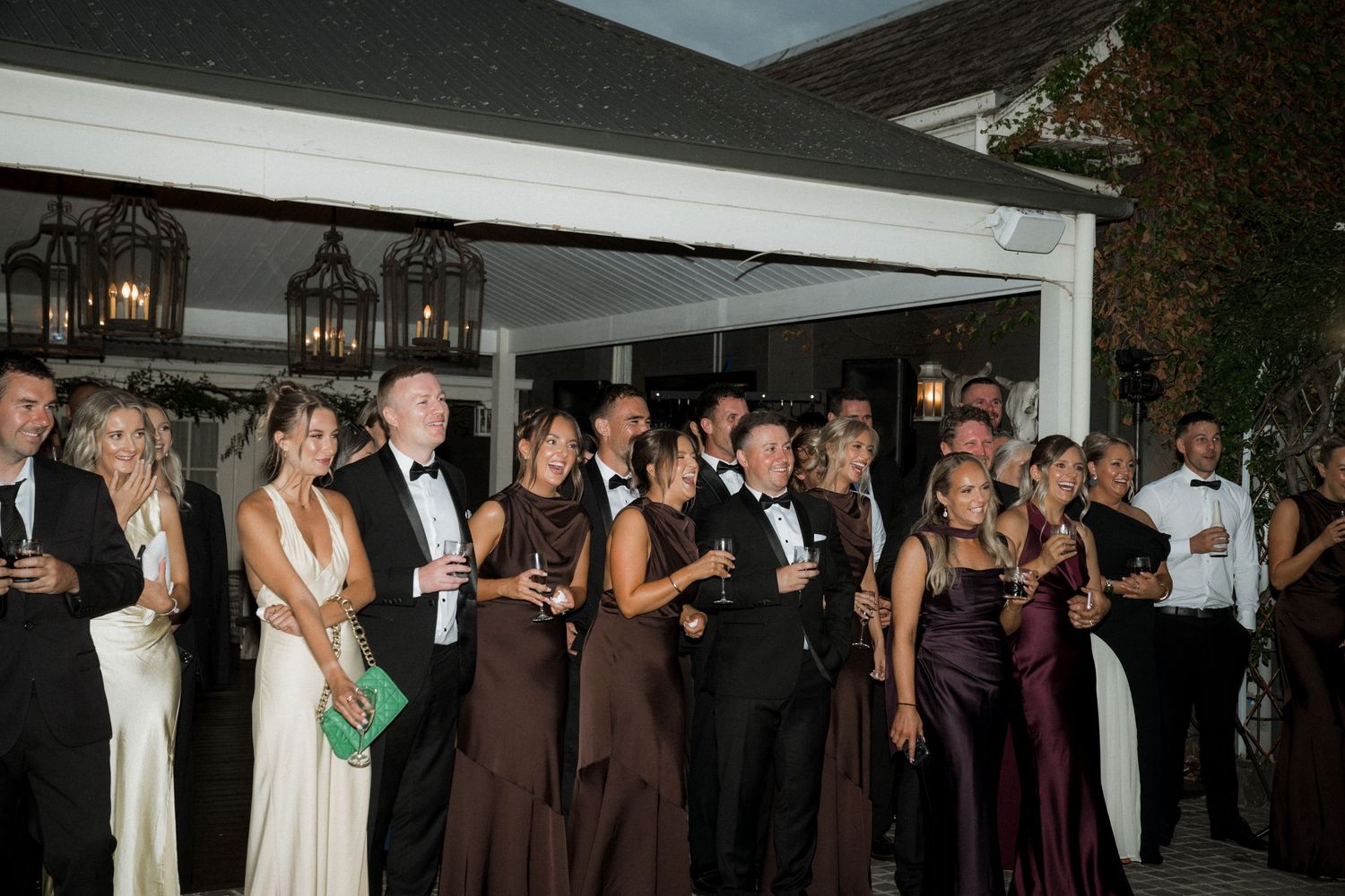 Elegant party guests in formal attire gather under a white canopy during an evening celebration.