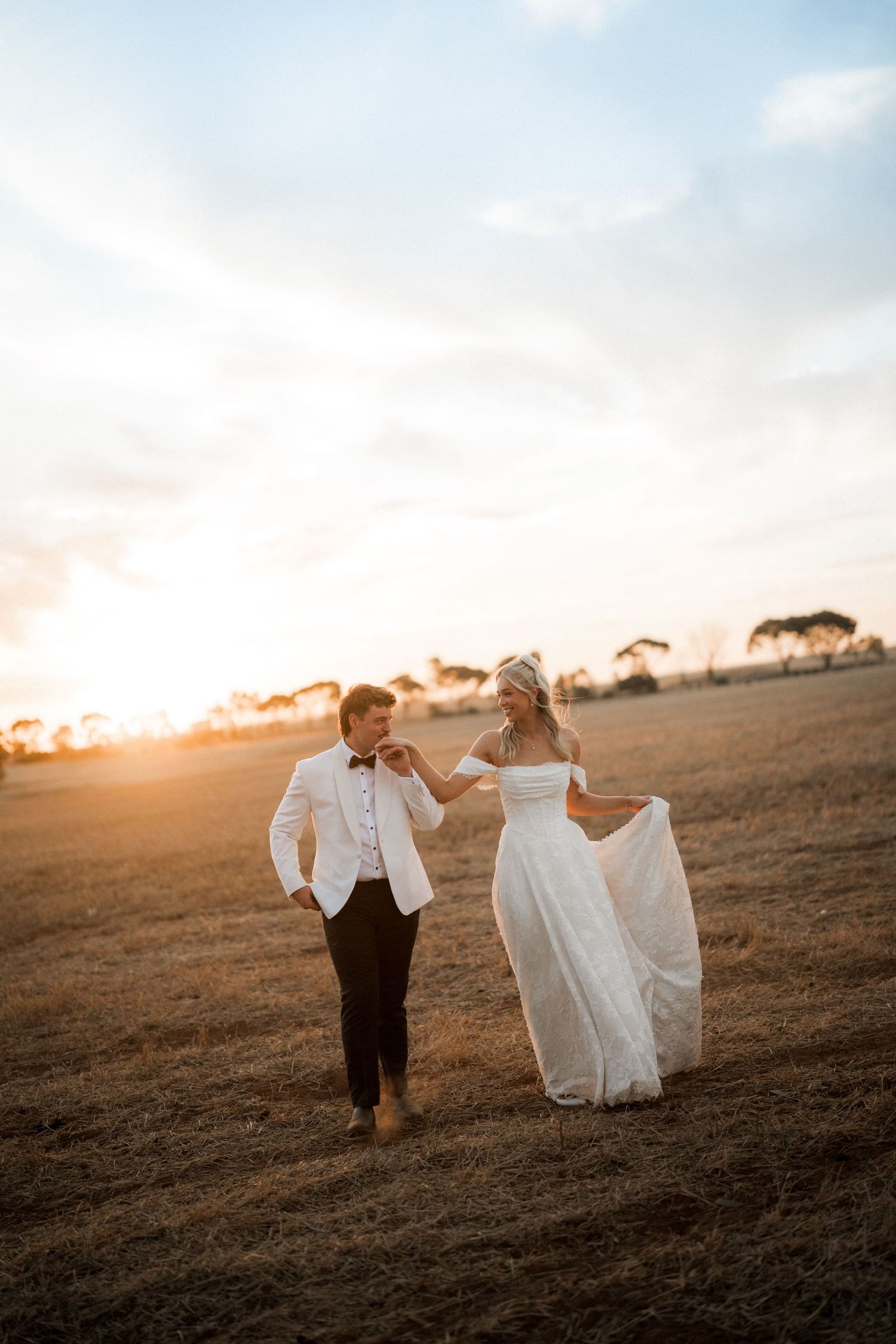 Bride and groom walk through golden field at sunset, bride holding her white gown.