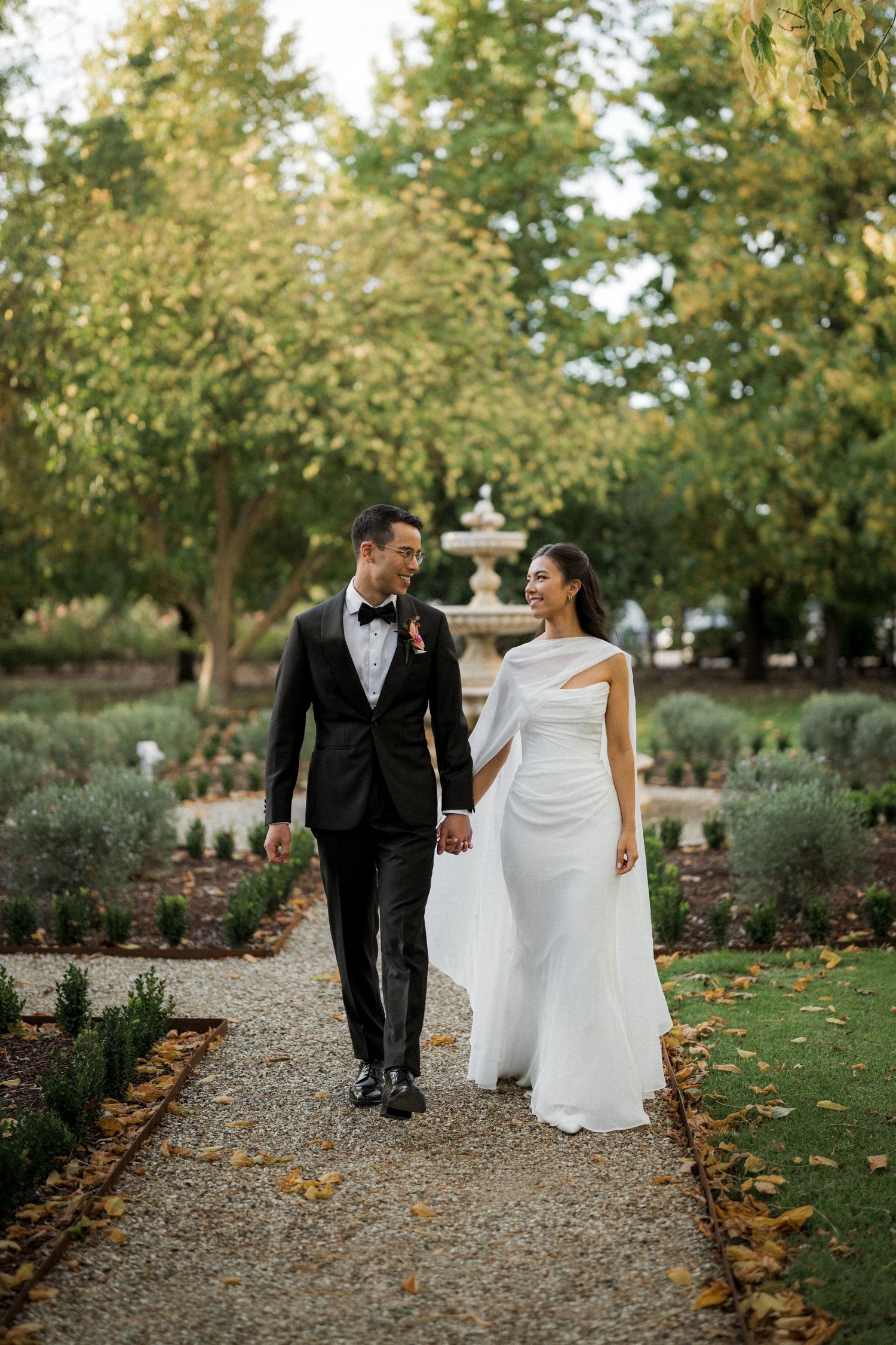 Bride and groom walking hand in hand along garden path with fountain and autumn trees.