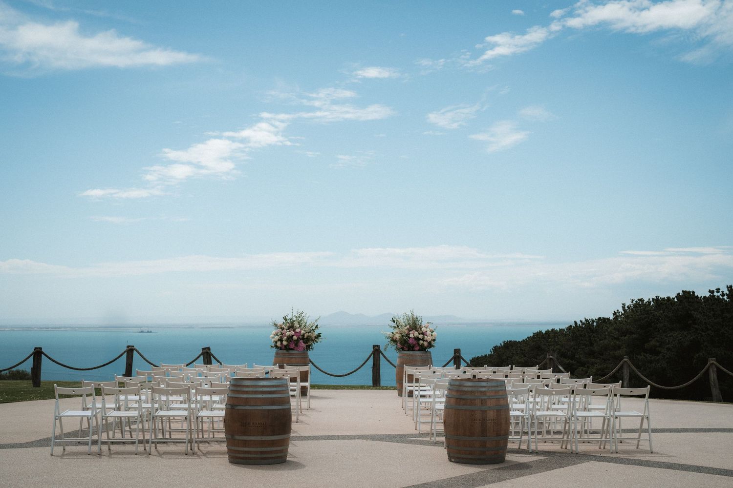 An elegant outdoor wedding ceremony setup with rustic wine barrels and white chairs overlooking the ocean.