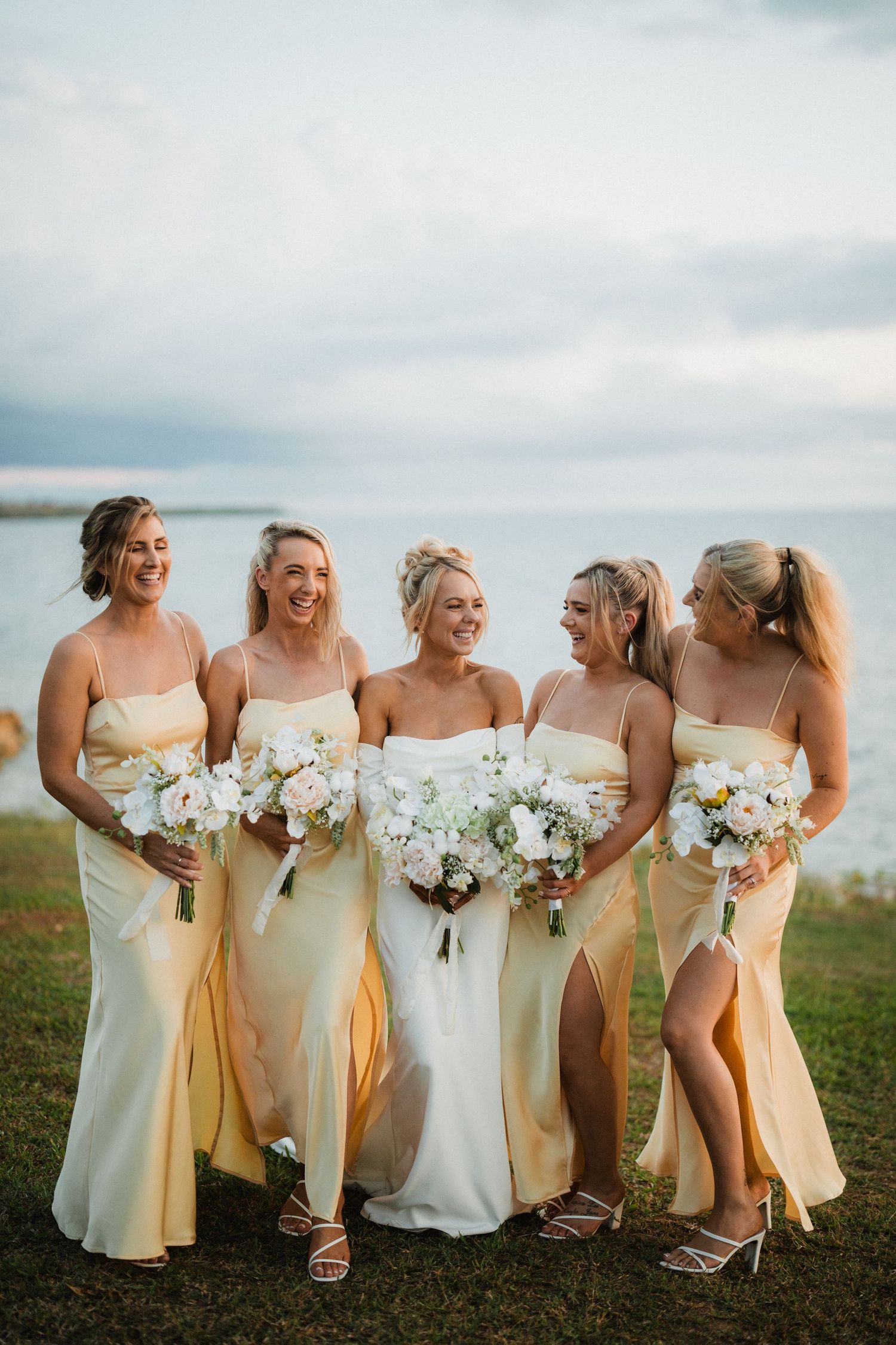 A bridal party in pale yellow dresses poses together on a coastal cliff while holding white floral bouquets.