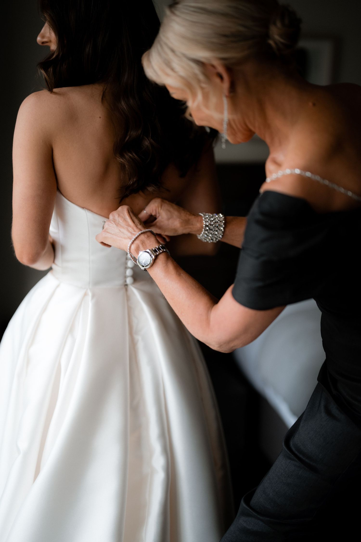 Helper adjusts back of white wedding dress while bride stands in intimate getting ready moment.