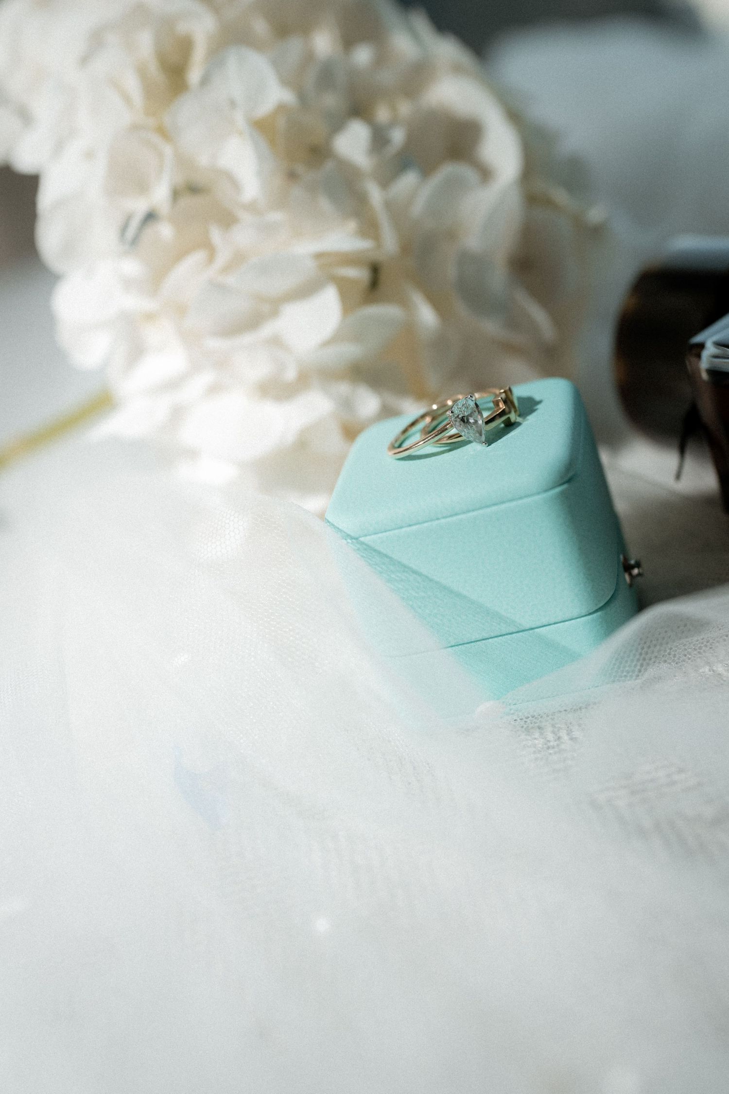 A diamond engagement ring sits in a mint green jewelry box next to white flowers on a soft textured surface.