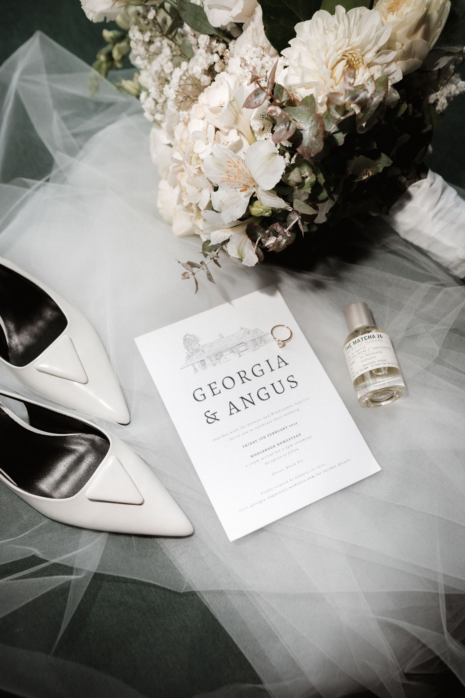 Wedding invitation and white shoes with floral arrangement and veil on dark surface.